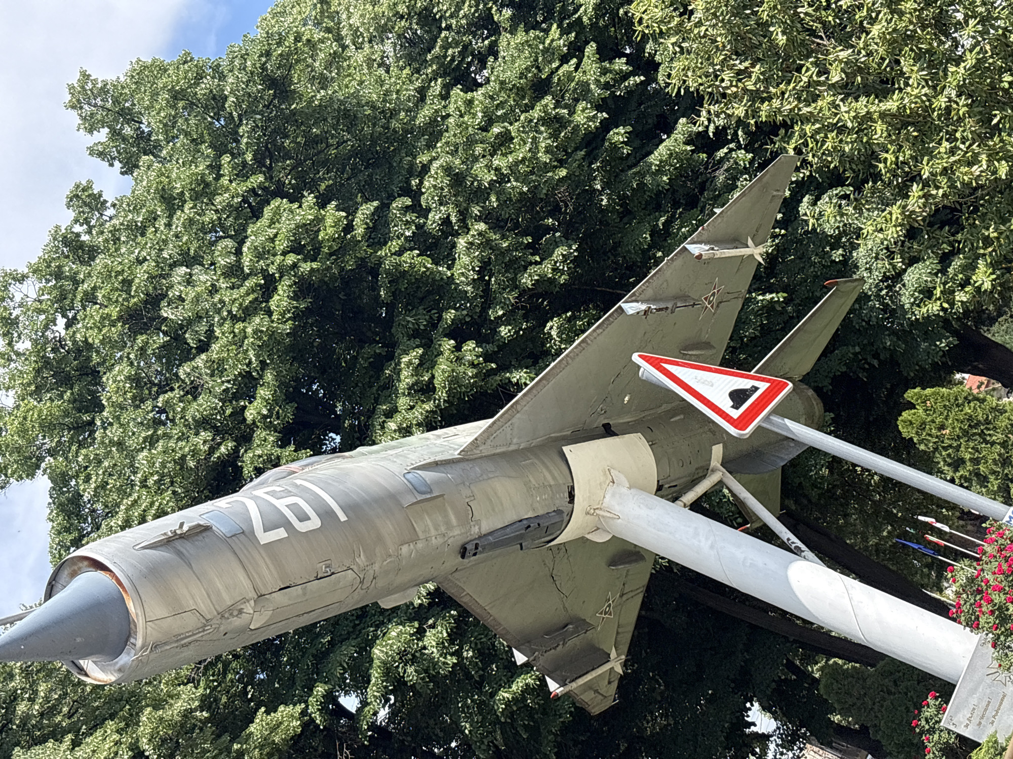 The image shows a Mikoyan-Gurevich MiG-21 fighter jet mounted on display in Yoakim Gruevo, Bulgaria. The aircraft is painted in a grey camouflage scheme with the number "261" visible on its fuselage. It is mounted on a metal support structure, with a road sign attached near the tail section. The background consists of dense green trees and vegetation. The jet appears to be in a public area or park, likely a memorial or monument.