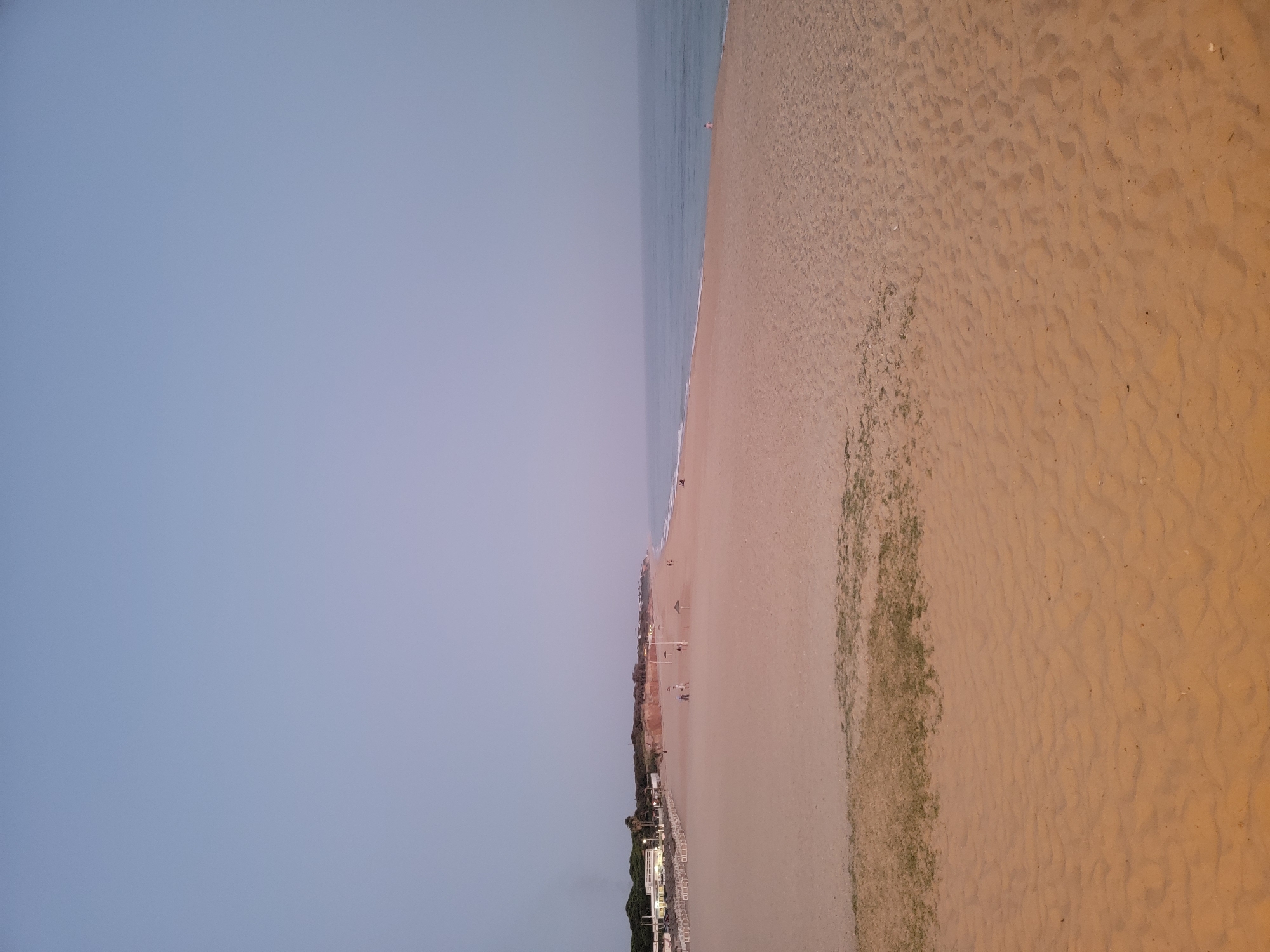 A wide, sandy beach stretches along the coast of Quarteira, Portugal, with the calm ocean to the right and buildings and vegetation to the left. The sky is a pale, hazy blue, suggesting either early morning or late evening light. The sand is a warm, golden-brown color, with subtle ripples and some patches of low-lying green vegetation. A line of beach chairs and umbrellas is visible in the distance on the left side of the beach, in front of white buildings that appear to be part of a resort or hotel. A few scattered figures are present on the beach, some walking and others seated. The ocean is a muted blue-grey, with small waves gently rolling onto the shore. The overall scene is serene and peaceful, with a sense of quietude.