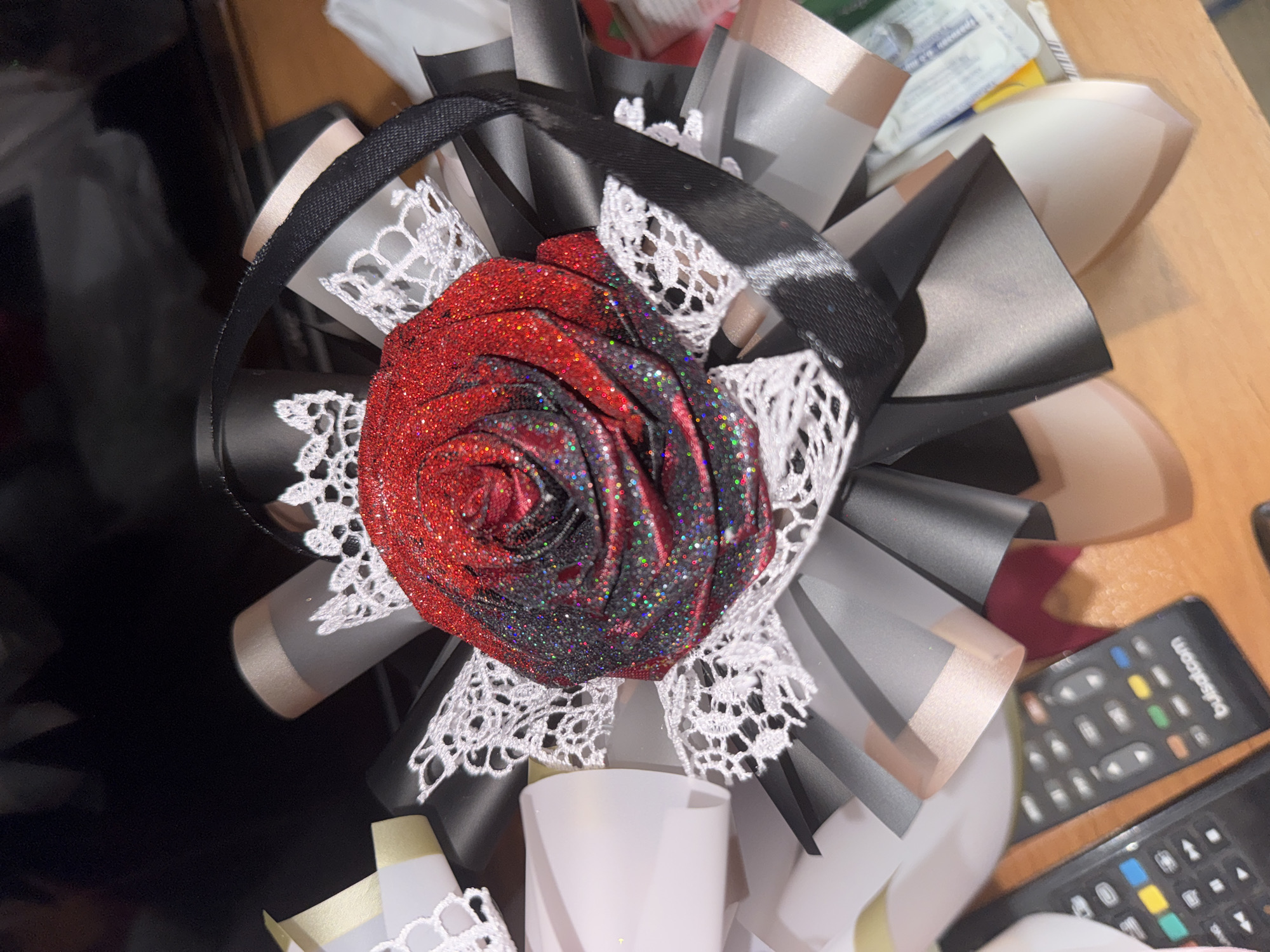 The image shows a close-up of a decorative arrangement, likely a bouquet or gift, featuring a large, glitter-covered rose in shades of red and black. The rose is surrounded by black and silver ribbons, white lace trim, and other decorative elements. In the foreground, two black remote controls are visible on a wooden surface. One remote has a label that reads "bulsatcom". The background is dark and out of focus, showing what appears to be a shelf or table. There is no indication of the city of Sredets, Bulgaria, in the image.