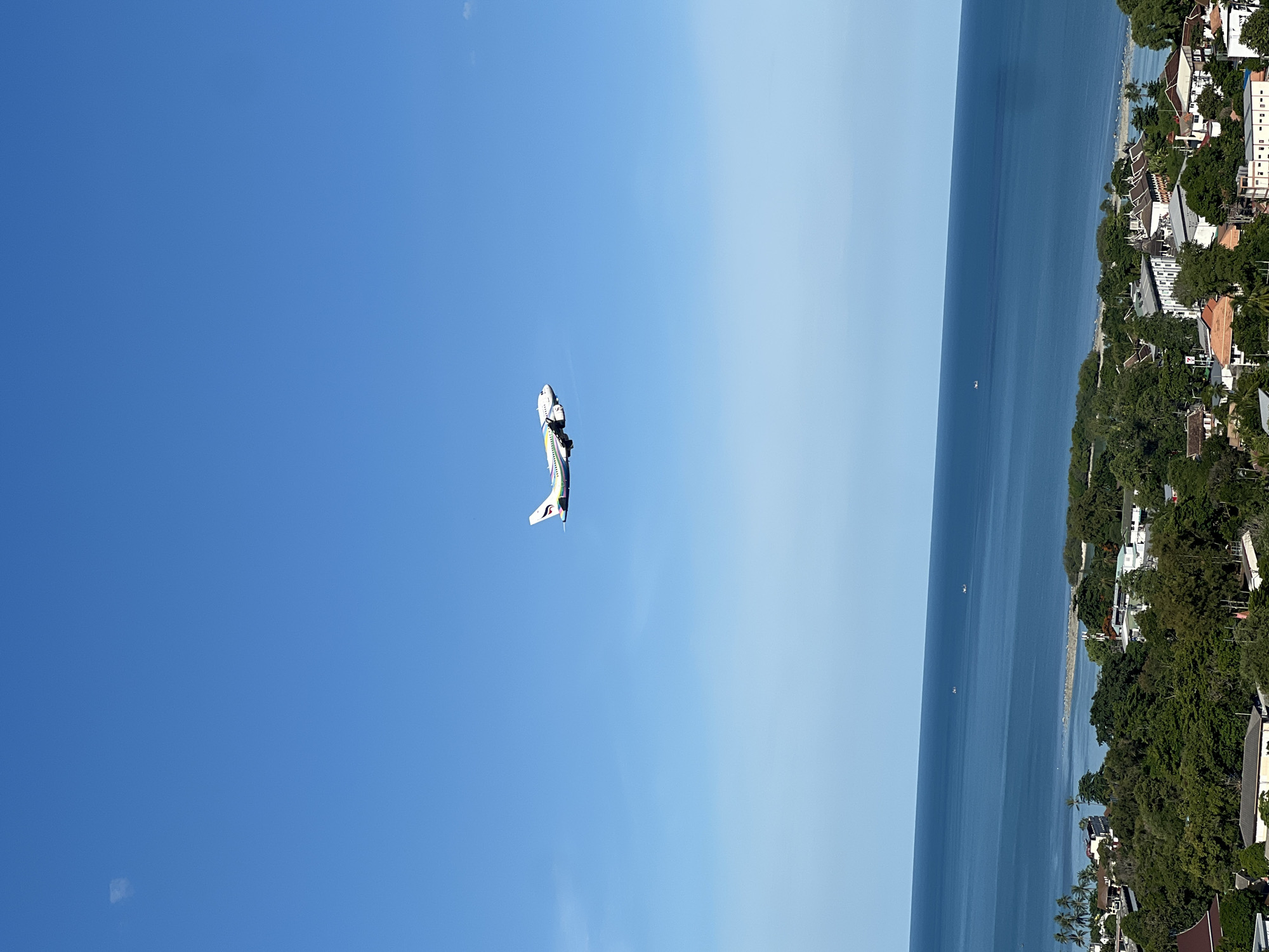 A prop plane is captured in mid-flight against a bright blue sky. The aircraft, predominantly white with green and red accents, is viewed from below, its underside visible. The plane appears to be a single-engine model, possibly a trainer or aerobatic aircraft, given its size and configuration.
The scene transitions to a serene coastal view. A vast expanse of calm, deep blue ocean stretches horizontally, meeting a distant, hazy horizon. Along the coastline, a lush green landscape is dotted with buildings. These appear to be houses or resort accommodations, with white walls and terracotta or dark-colored roofs, nestled amongst dense foliage. Some palm trees are visible, suggesting a tropical or subtropical climate. The water near the shore is lighter blue, and a sandy beach is visible in places. The overall impression is one of a clear, sunny day in a tropical location.
The orientation of the image is such that the sky is on the left and the ocean and land are on the right. There is no visible text on the plane or in the landscape. The time of day is clearly daytime, with bright sunlight casting minimal shadows. The weather appears to be fair and calm, ideal for both flying and enjoying a coastal environment. The overall mood is peaceful and tranquil.