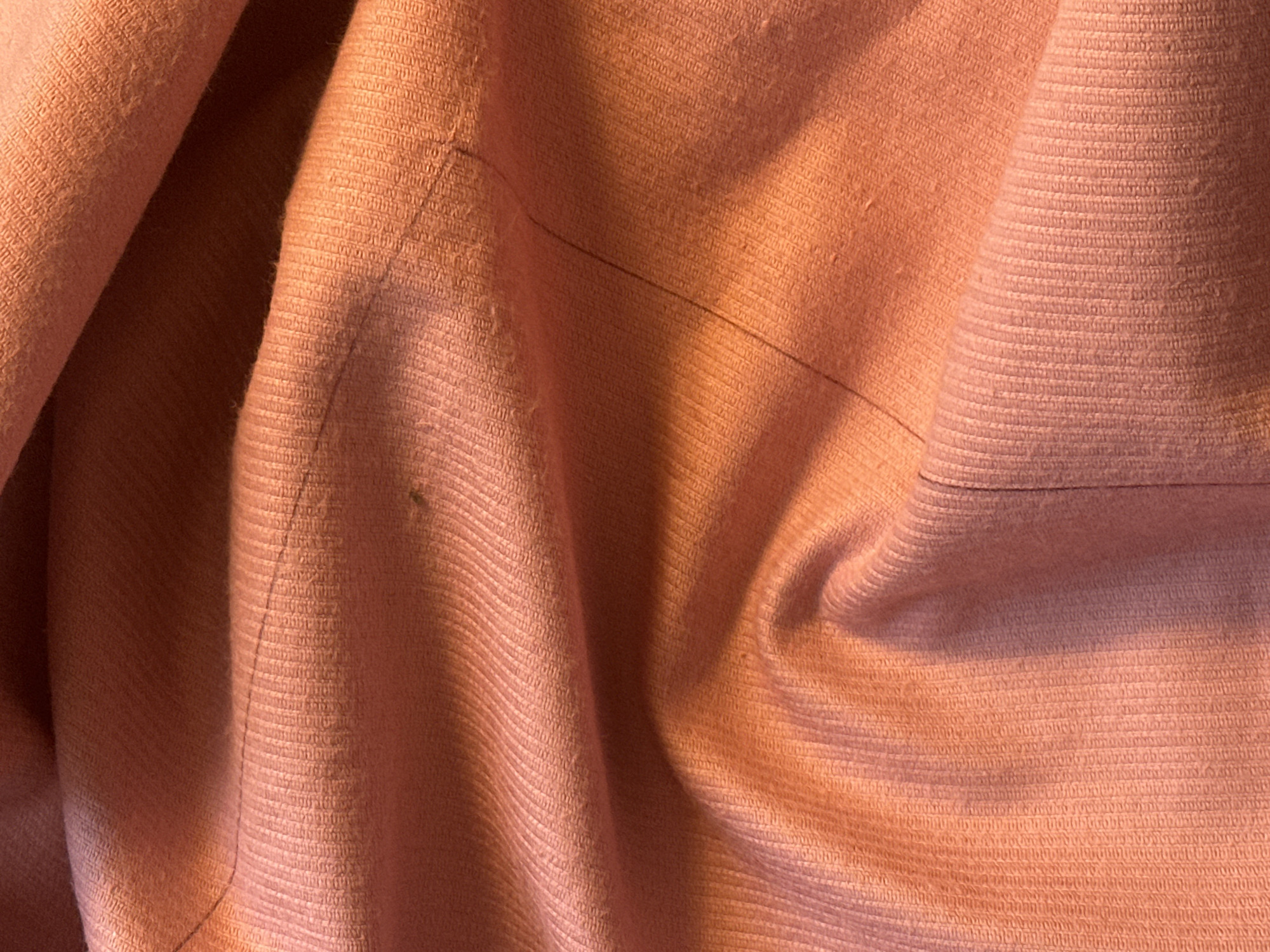 The image shows a close-up of a crumpled piece of peach-colored fabric. The texture is visible, with a woven pattern. There are lines or creases running across the fabric, and a small spot or imperfection is noticeable on the left side. The lighting is warm, creating shadows and highlights that emphasize the folds and textures of the fabric. The scene has no discernible location in Buenos Aires, Argentina, and contains no information about people, subjects, objects, actions, interactions, or the environment beyond the fabric itself.