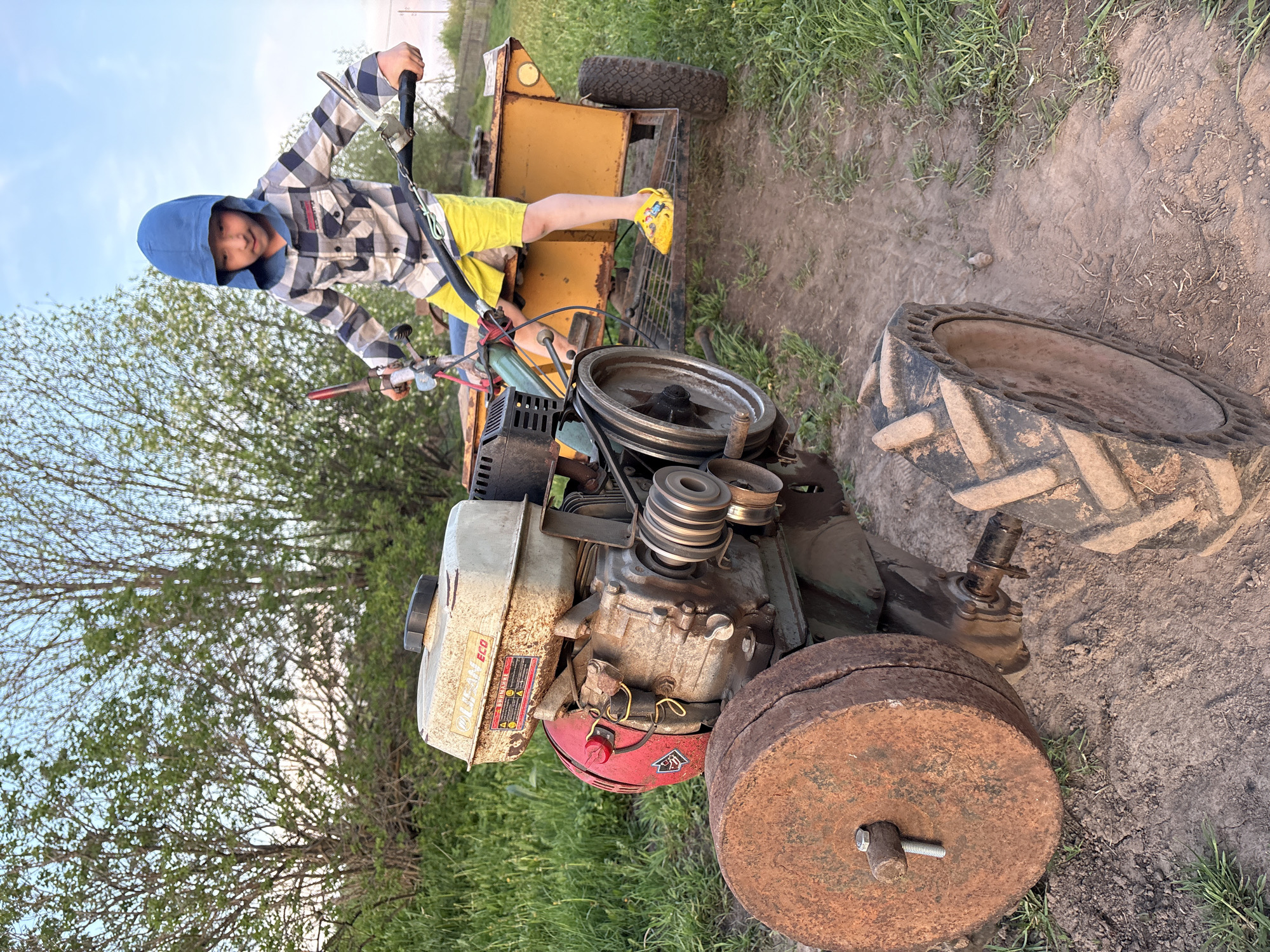 A child is sitting on a yellow tractor in an outdoor setting. The child is wearing a blue hooded sweatshirt and yellow shorts. The tractor is old and rusty, with a visible engine and wheels of varying sizes. The ground is dirt and grass. Trees are visible in the background. The location is Nizhnyaya Chermoda, Russia.