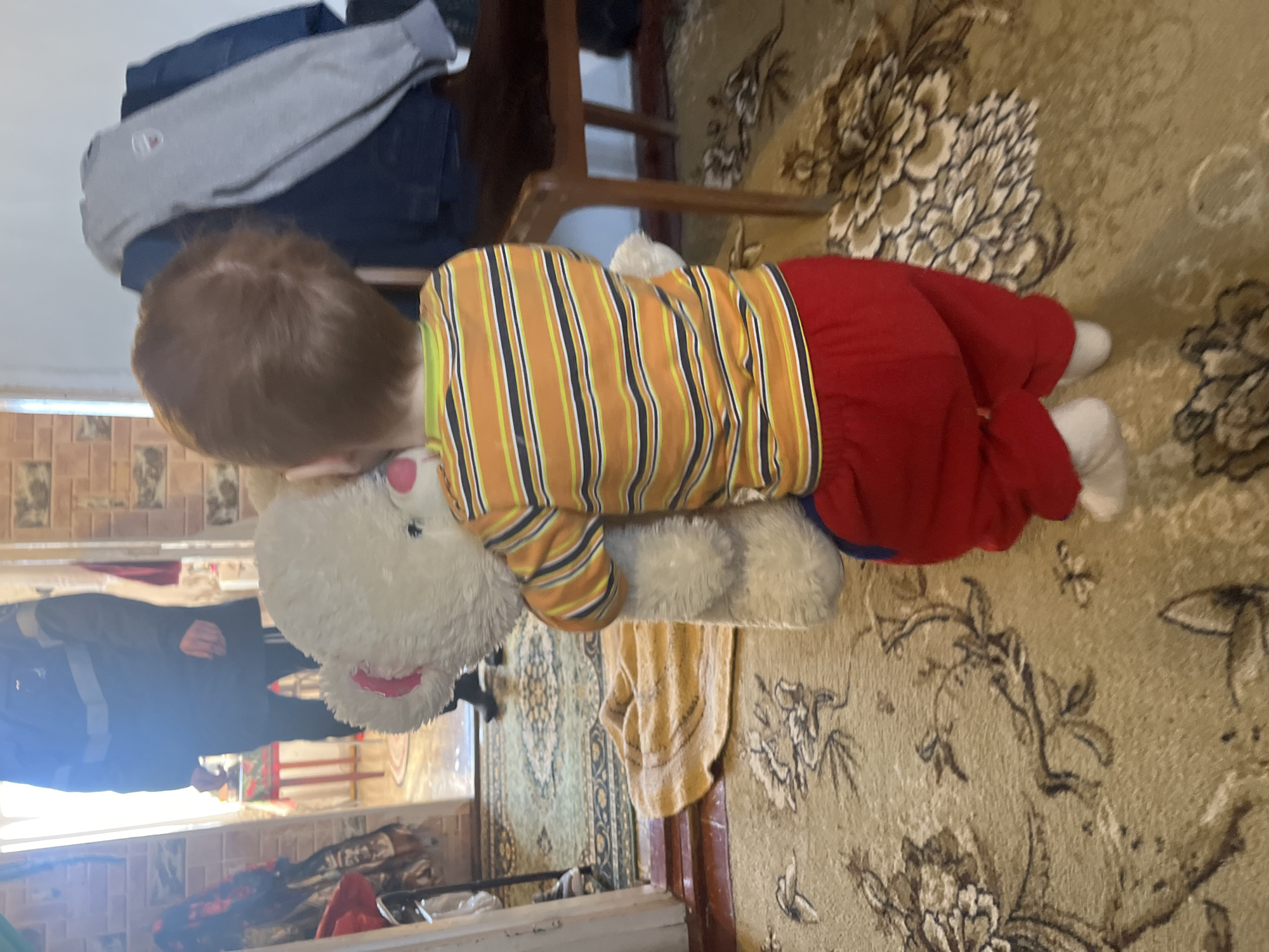 A child with red hair is lying on a tan rug with a floral pattern in an indoor setting. The child is wearing a striped yellow and orange shirt with red pants and white socks, embracing a large white stuffed animal. A wooden chair with clothing items on top is visible in the background. A person in dark clothing is visible in the reflection of a window. Based on the location provided, the image was taken in Сосново, Russia.