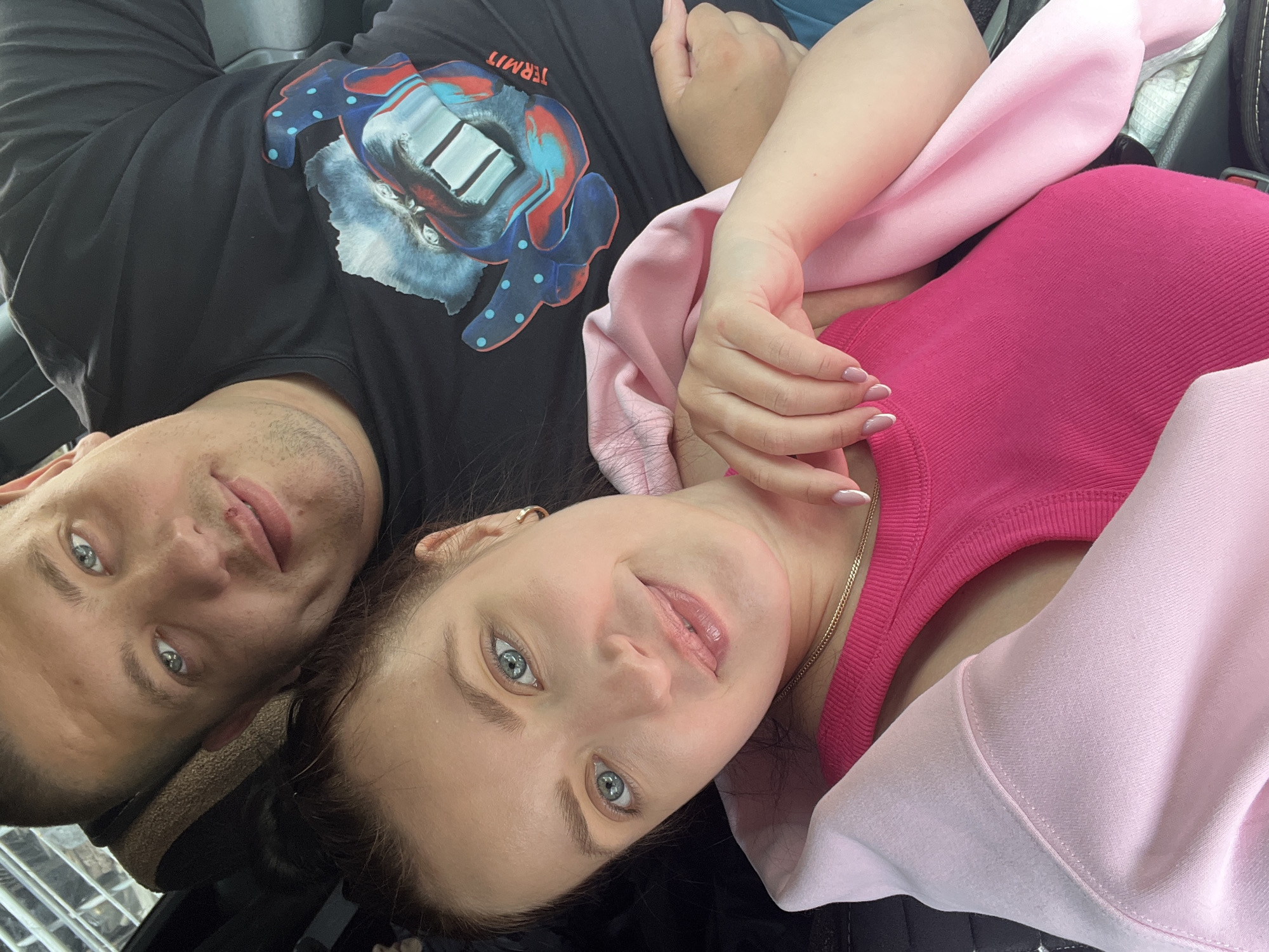 Two people, a man and a woman, are lying down in close proximity. The man is wearing a black t-shirt with a colorful graphic on it. The woman is wearing a pink top and a light pink hoodie. They are both looking towards the camera. The background is unclear, possibly a car interior. The location is not discernible as Chaikovsky, Russia, based on the visual information provided.