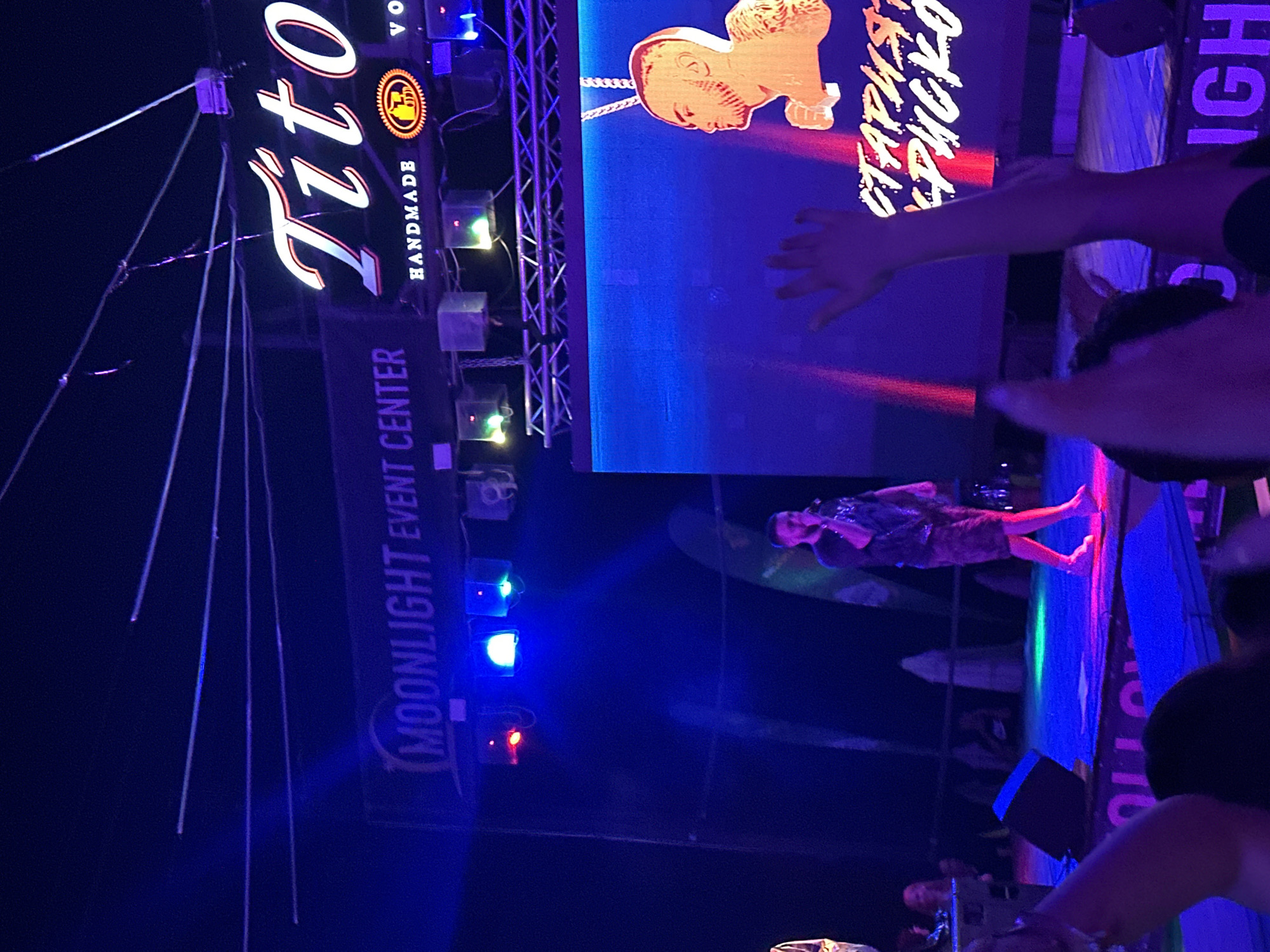 The image shows a concert or event taking place at the Moonlight Event Center in Varna, Bulgaria. A male performer is on stage, holding a microphone and singing. He is wearing a dark t-shirt and shorts. Behind him, a large screen displays a graphic of a man's head with a gold chain. The stage is lit with blue and pink spotlights. In the foreground, audience members' hands are raised, suggesting they are enjoying the performance. The "Tito's Handmade Vodka" logo is visible above the stage. The overall atmosphere appears to be energetic and festive.