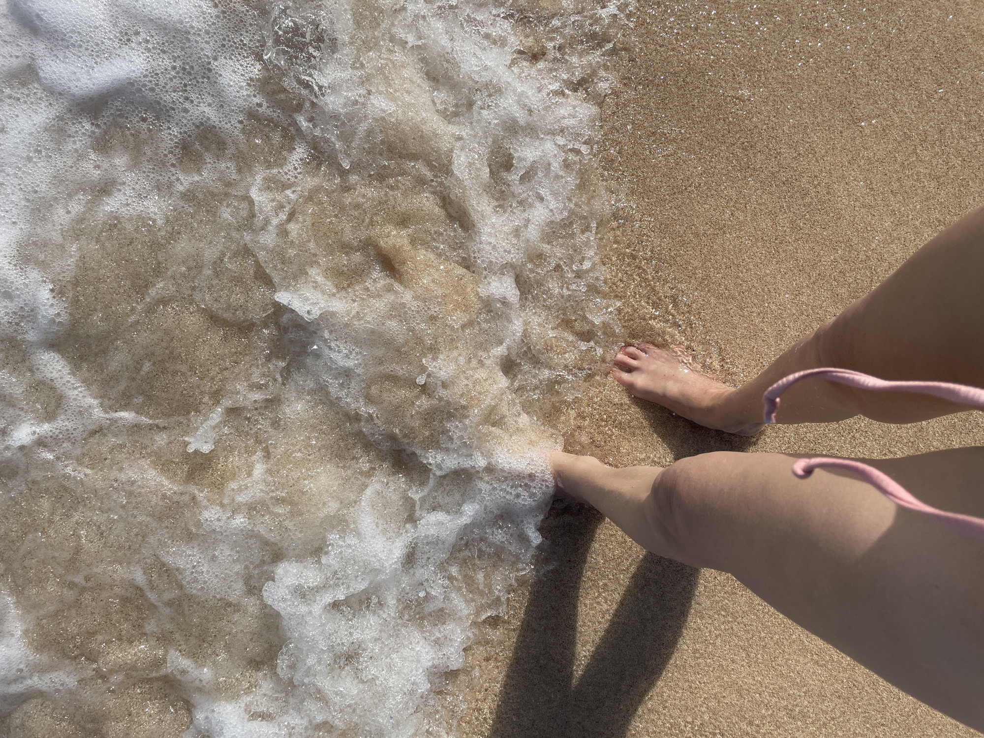 A first-person perspective shot captures a person's legs and feet standing on a sandy beach, with a gentle wave of white foam washing over their feet and toes. The sand is a light tan color and appears wet and glistening where the water has receded. The water itself is clear, with visible sand grains underneath, and it forms frothy bubbles as it interacts with the shore. A prominent shadow, cast by an unseen object, stretches across the sand from the left side of the frame. The person is wearing a pink bikini. The location is identified as Kuta Selatan, Indonesia.