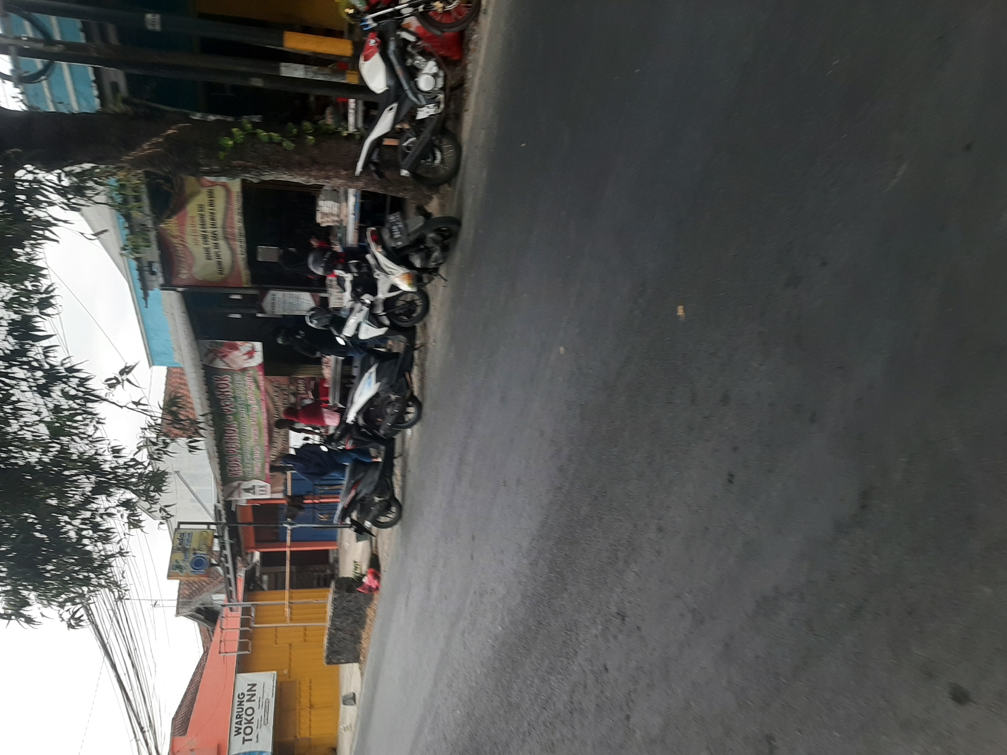 The image shows a street scene in Sumampir, Indonesia, with several motorcycles parked along the side of the road. In the background, there are various shops and buildings. One shop has a sign that reads "WARUNG TOKO NN". Another shop has a banner with Indonesian text. There are a few people visible, some standing near the motorcycles and others inside the shops. The environment appears to be an urban or semi-urban area with a paved road and sidewalks. Power lines are visible overhead.