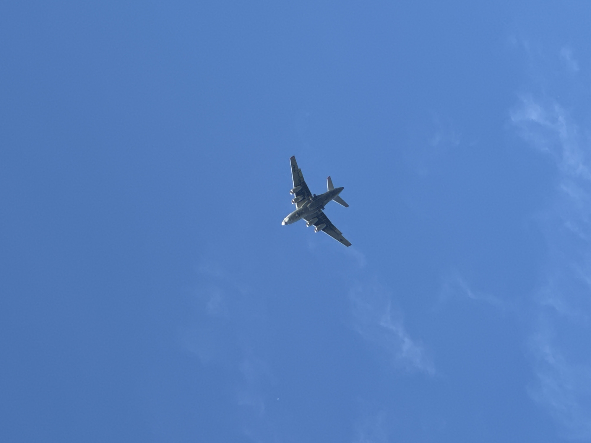 A military transport aircraft, possibly an Antonov An-12, is captured mid-flight against a clear, bright blue sky. The aircraft is oriented with its nose pointing upwards and to the right, viewed from below. Its four turboprop engines are visible under its high-wing configuration, along with its distinctive T-tail. The aircraft appears to be camouflaged with a mottled pattern.
The sky is predominantly clear, with only wisps of thin, high-altitude clouds scattered sparsely, suggesting a daytime scene with good visibility. The lighting indicates it is likely daytime, perhaps mid-morning or afternoon, given the brightness and lack of deep shadows. The perspective from below the aircraft suggests the photo was taken from the ground, looking directly up.
No people are visible in the frame. The scene is solely focused on the aircraft in flight against the vast expanse of the sky. There are no discernible location cues beyond the clear sky, and no text is visible on the aircraft or in the environment. The overall impression is one of an operational flight in fair weather conditions.