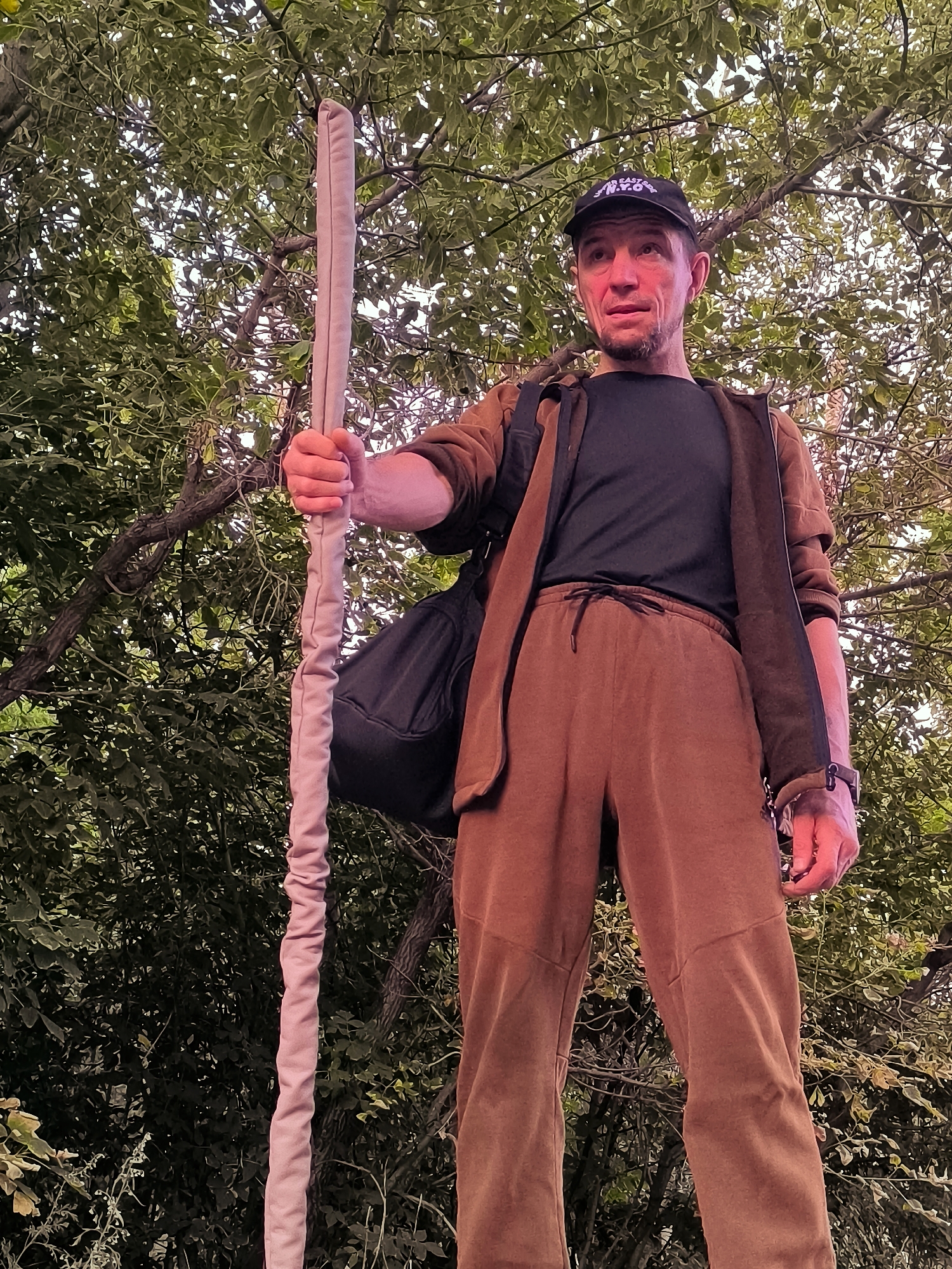 A low-angle full shot shows a man holding a staff in front of a backdrop of trees and foliage. He is wearing a black baseball cap, a dark t-shirt, a brown zip-up jacket, and brown sweatpants. A black bag is slung over his left shoulder. His skin is illuminated with a reddish hue, possibly from the lighting. The man appears to be standing outdoors. The scene does not provide any specific indicators to place it within the city administration of Qarağandı, Kazakhstan.