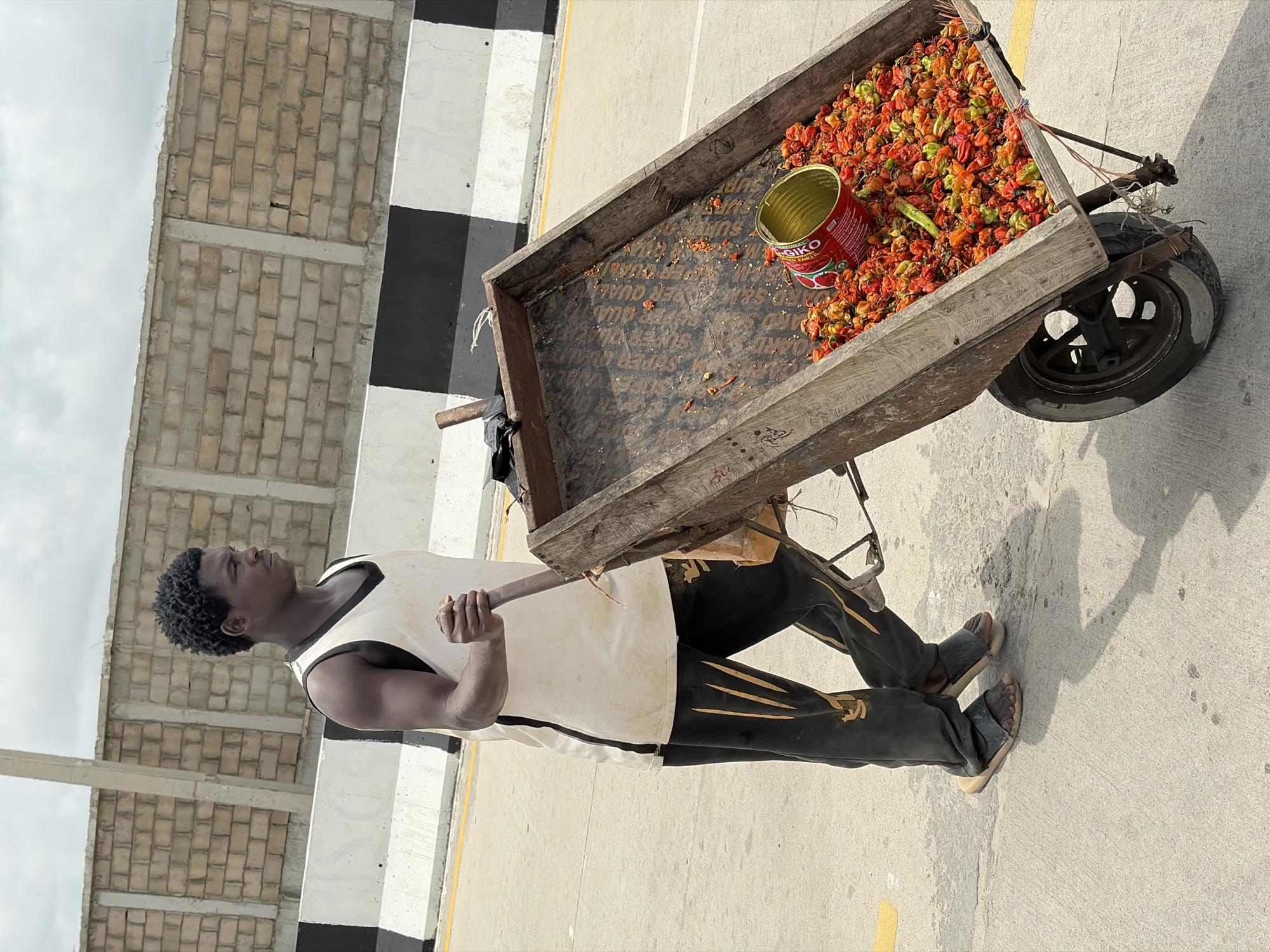 A man of African descent is seen pulling a cart filled with red peppers in Igboefon, Nigeria. The cart is constructed of wood and has a single wheel. A red can is present among the peppers. The man is wearing a white tank top, dark pants with light markings, and sandals. He has short, dark hair and is looking towards the left of the frame. The background features a brick wall with alternating black and white sections. The setting appears to be an urban or roadside environment.
