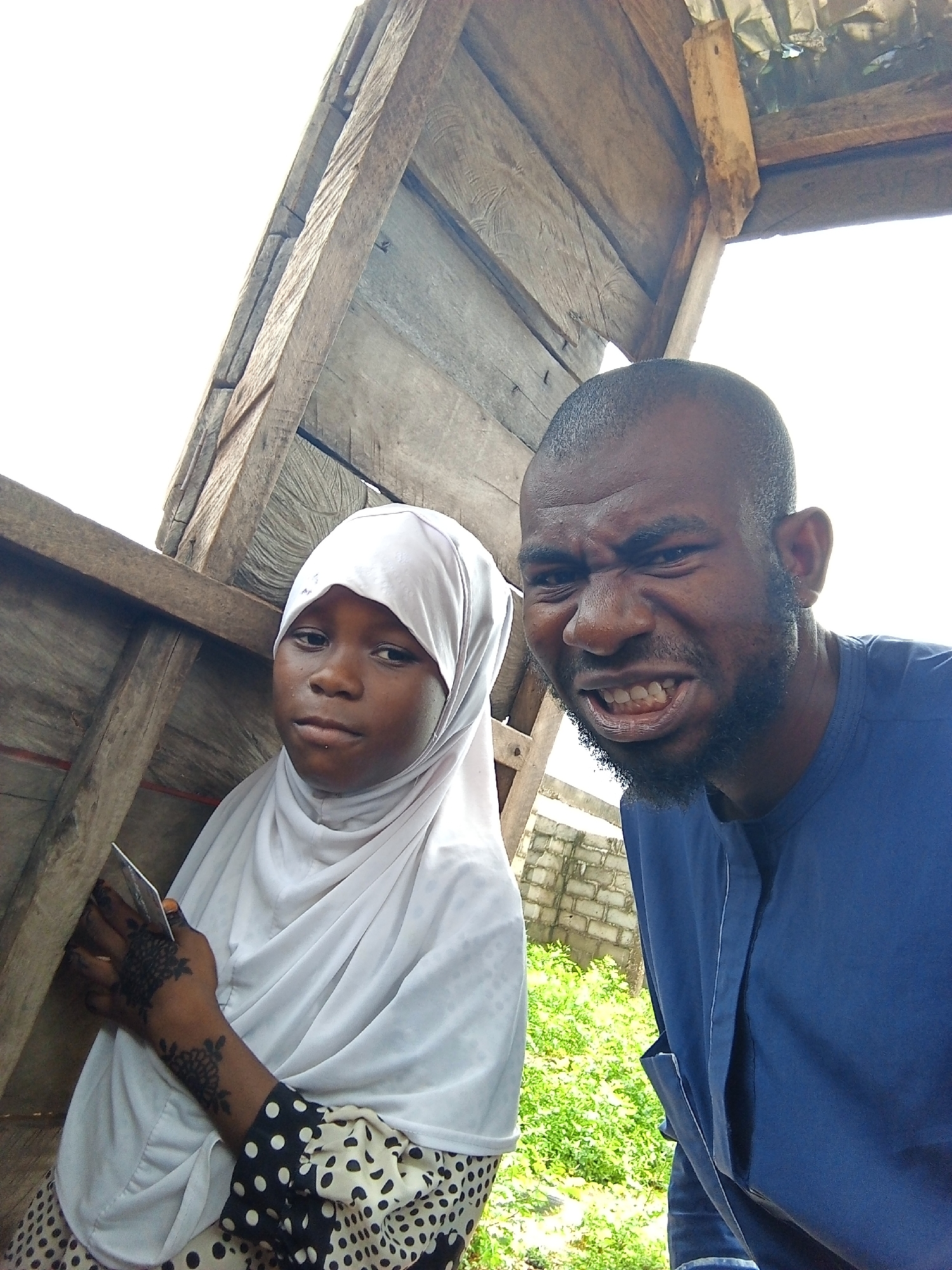 The image depicts two individuals under a wooden structure in an outdoor setting. A young woman wearing a white hijab and a polka-dotted top holds a card in her left hand, which is adorned with dark floral patterns. Next to her stands a man in a blue shirt, grimacing at the camera. The environment includes glimpses of greenery and a brick structure in the background, suggesting an outdoor location within an urban area. The wooden structure overhead is made of rough planks and beams, possibly a makeshift shelter or part of a building under construction.