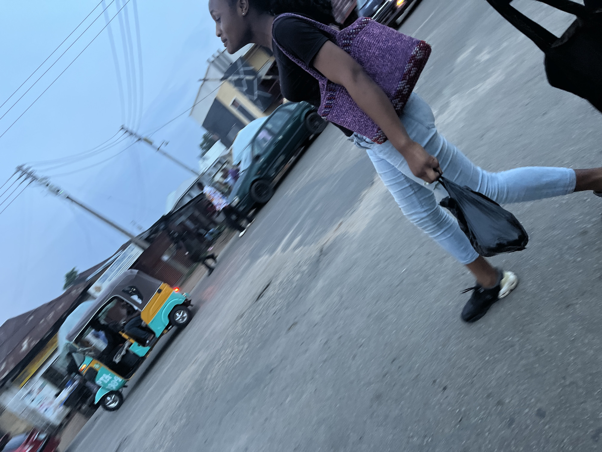 An outdoor, eye-level, medium shot shows a young Black woman walking on a paved road. The woman is facing to the left and looking upwards. She is wearing a black t-shirt, light blue jeans, and black shoes. She is carrying a purple woven bag on her right shoulder and holding a black plastic bag in her left hand. A tuk-tuk is visible to the left of the woman, as is a green car further back. The scene appears to be a street in Jos, Nigeria, with a cloudy sky and utility poles visible in the background. The image is blurred due to motion.