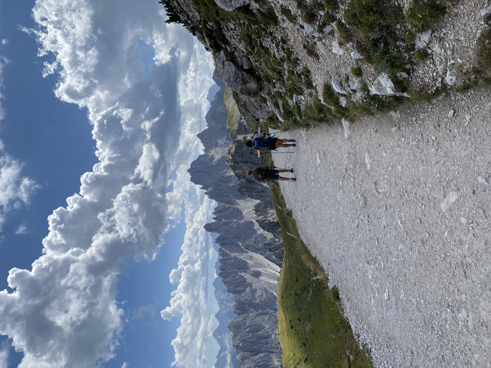 The image depicts a scenic mountain landscape in the vicinity of Auronzo di Cadore, Italy. A gravel path winds through the foreground, with two hikers, a man and a woman, walking away from the viewer. The man is on the left, wearing a hat and shorts, and carrying a backpack and trekking poles. The woman is to his right, with a backpack and trekking poles. The path is bordered by sparse vegetation and rocky terrain on the right, and a grassy slope with a distant fence on the left. In the background, jagged, imposing mountain peaks dominate the horizon under a sky filled with dramatic, billowy clouds. The overall scene conveys a sense of remote natural beauty and outdoor activity.