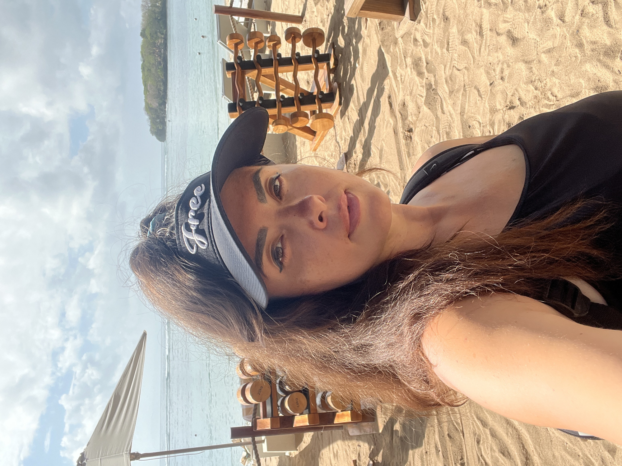 A medium shot captures a woman in a black visor with the word "Free" embroidered in white, facing the camera outdoors on a sandy beach. She has long, reddish-brown hair pulled into a ponytail and is wearing a black tank top. The background reveals a beach setting with a calm ocean under a partly cloudy sky. To her left, a beige beach umbrella is partially visible. In the mid-ground, two wooden racks hold dumbbells, with the weights on the left rack labeled with numbers and "kg." Beach chairs and wooden posts are also present. The sand is imprinted with footprints. The scene is likely at a resort or gym facility located near the ocean in Kuta Selatan, Indonesia, given the presence of fitness equipment on the beach.