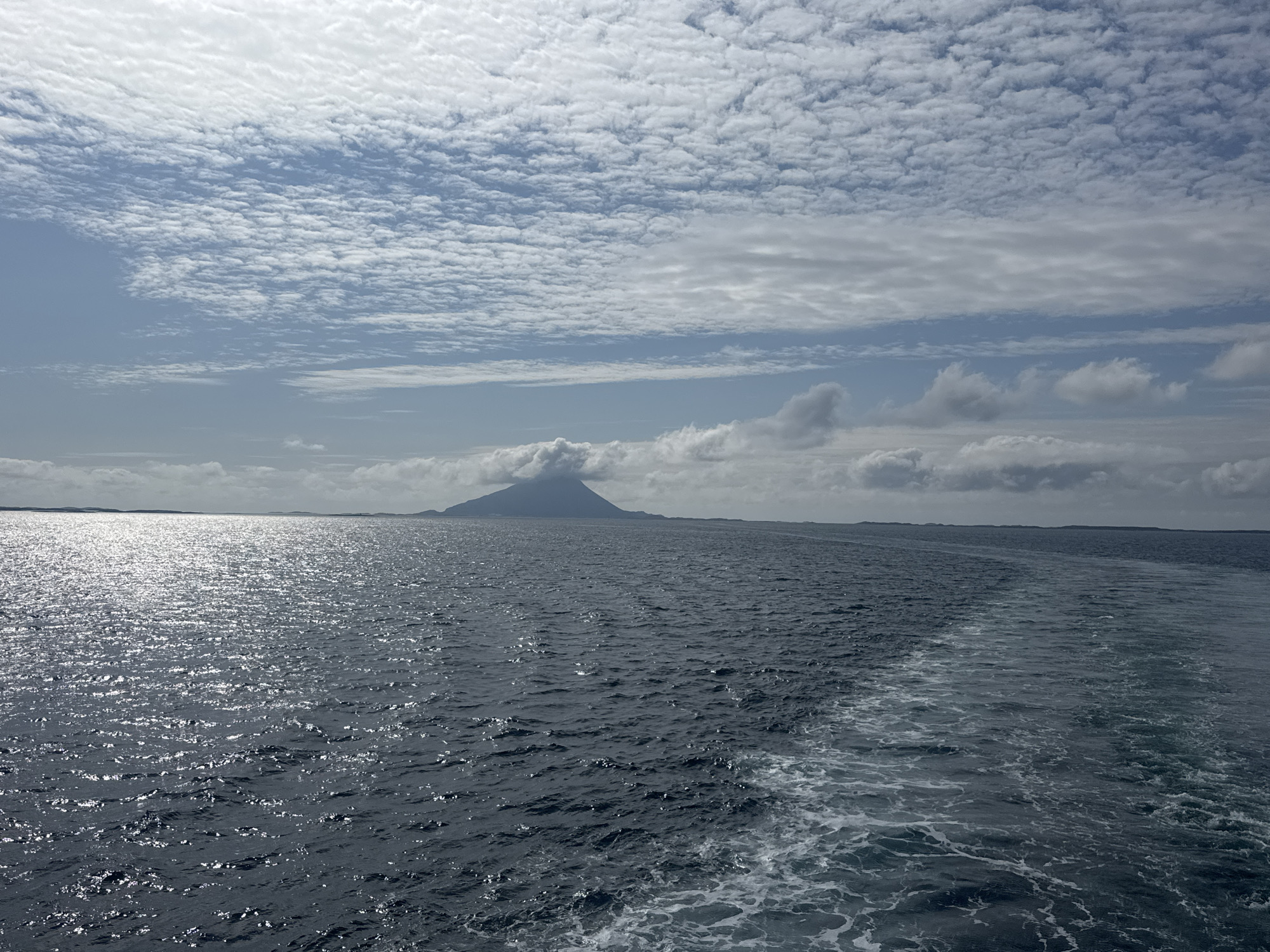 The image depicts a vast expanse of ocean under a partially cloudy sky. In the distance, a conical mountain rises from the sea, its peak shrouded in clouds. The water is dark blue with shimmering reflections of sunlight on its surface. A foamy wake trails behind the viewer, indicating movement through the water. The horizon is marked by a faint outline of land to the left and right of the mountain. The scene is serene and natural, with no human presence or man-made structures immediately visible. The specific location in the city of Sleneset, Norway, cannot be determined from the image alone.