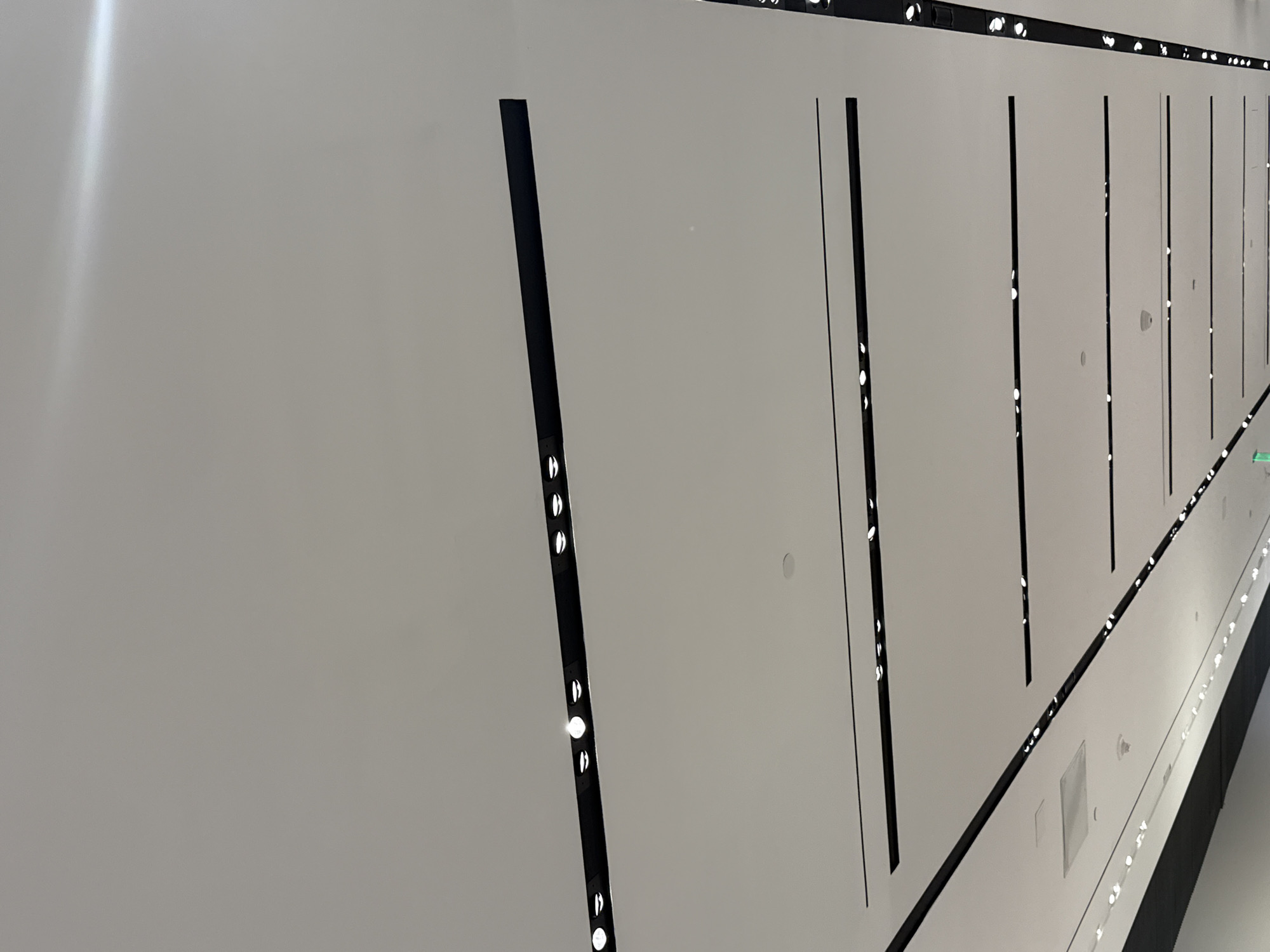 The image depicts a minimalist, modern interior architectural space, likely a corridor or gallery, characterized by clean lines and uniform lighting. The dominant feature is a long, light grey or off-white wall that stretches into the distance, suggesting a significant length.
This wall is punctuated by regularly spaced vertical black strips, which act as architectural accents and house numerous small, circular, bright white LED lights embedded within them. These lights illuminate the space and create a repetitive, rhythmic pattern along the wall. A similar black strip with embedded lights runs horizontally along what appears to be the top edge of the wall, possibly where it meets the ceiling, and another along the bottom where it meets the floor or a base structure.
No people are visible, and there is no discernible activity taking place. The scene is static and devoid of movement. The artificial lighting suggests an indoor setting where the time of day is not a factor. In the far distance along the corridor, a faint green glow or sign can be seen, hinting at further features or exits. Subtle circular fixtures or indentations are also present on the light-colored wall panels themselves. Towards the bottom right, a darker floor or base is visible, along with some rectangular panels or vents on the lower section of the wall. The overall impression is one of contemporary design, sleekness, and order, consistent with a modern public or commercial building in a location such as Miami, USA. No visible text is present in the image.