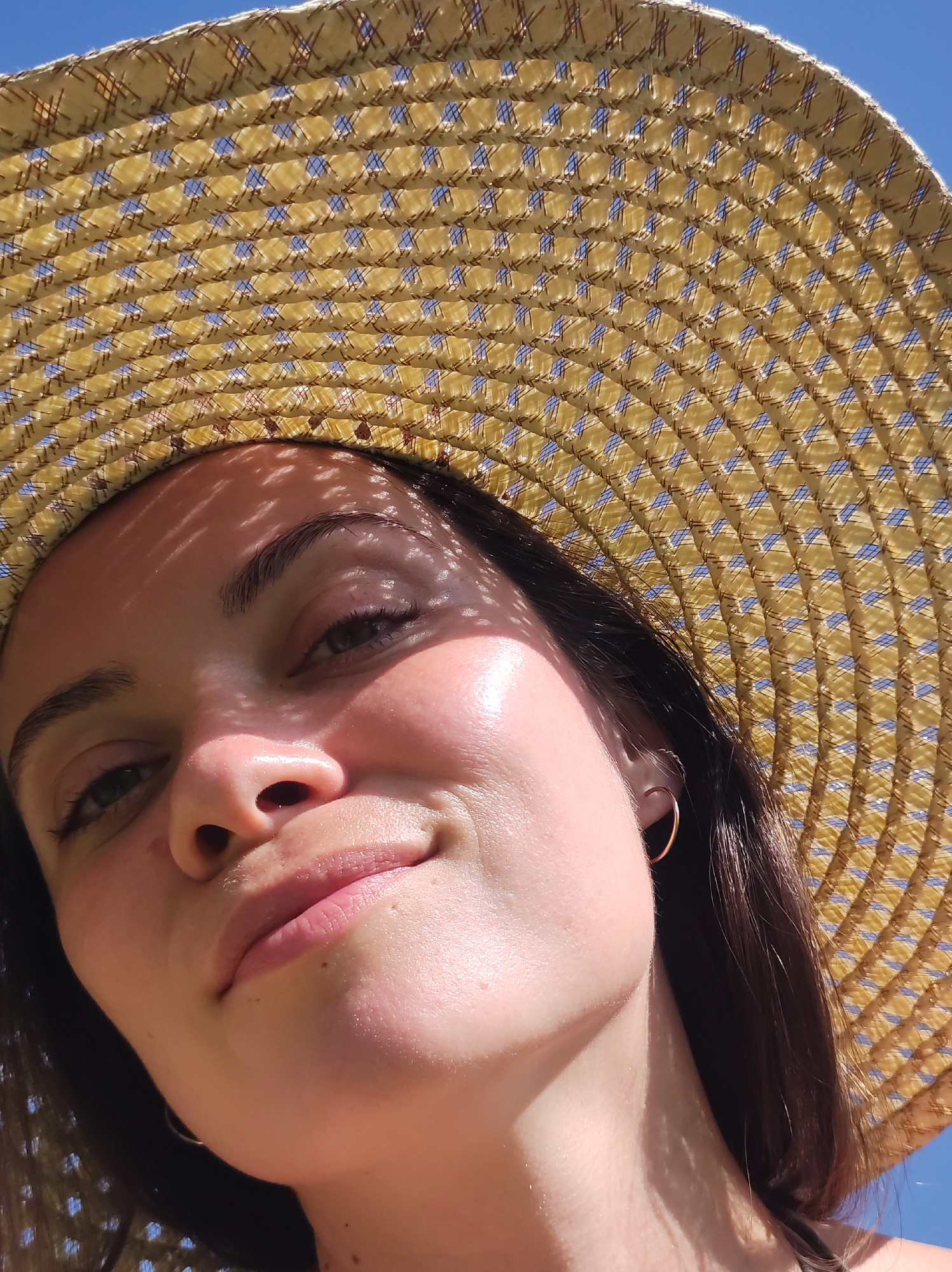 A woman is depicted from a low angle, wearing a straw sunhat. The hat creates a pattern of shadows on her face. She has dark hair, a gold hoop earring in her left ear, and fair skin. The background reveals a blue sky. The location is not discernible from this image.