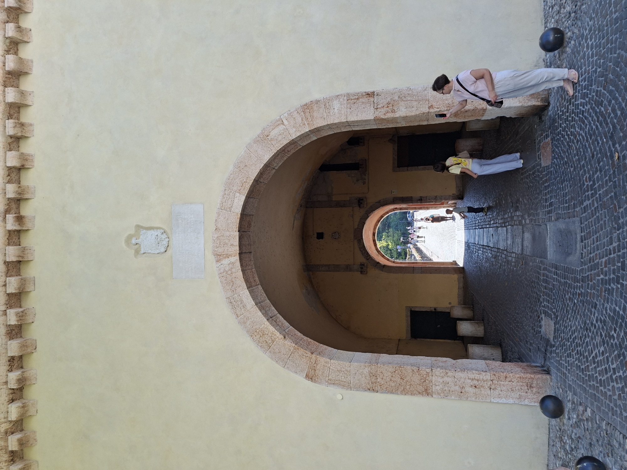 The image shows a wide, arched entryway into a building or fortification in Peschiera del Garda, Italy. The entryway is constructed of large stone blocks, and the wall above it is a pale yellow color, with decorative stone corbels along the top. A crest is visible on the wall above the arch, as is a rectangular plaque. Inside the arched entryway, another smaller archway is visible, leading to an outdoor area with a cobblestone path and several people. Two women and a child are in the foreground of the image, standing on a cobblestone area. One woman is taking a photo with her phone, and the other appears to be looking at her phone. A young boy stands near them, looking into the arched entryway. Two large, dark, spherical objects are on the ground in the foreground. The light suggests it is a sunny day.