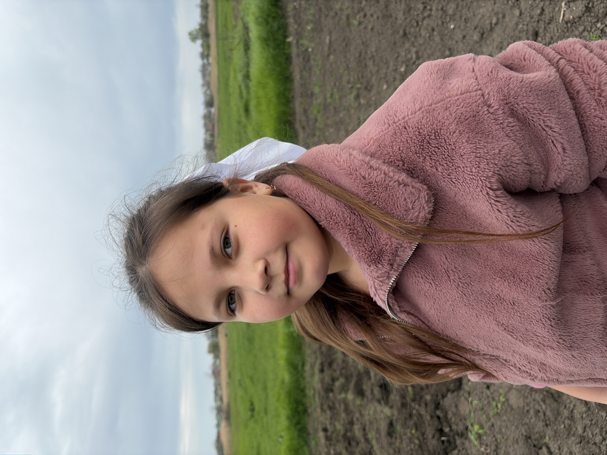 A young girl with brown hair and fair skin is outdoors in what appears to be a rural environment. She is wearing a mauve-colored, plush zip-up jacket and a white hair bow. The background features a green field, an area of brown dirt, and an overcast sky. The picture may have been taken in Novopokrovskaya, Russia.