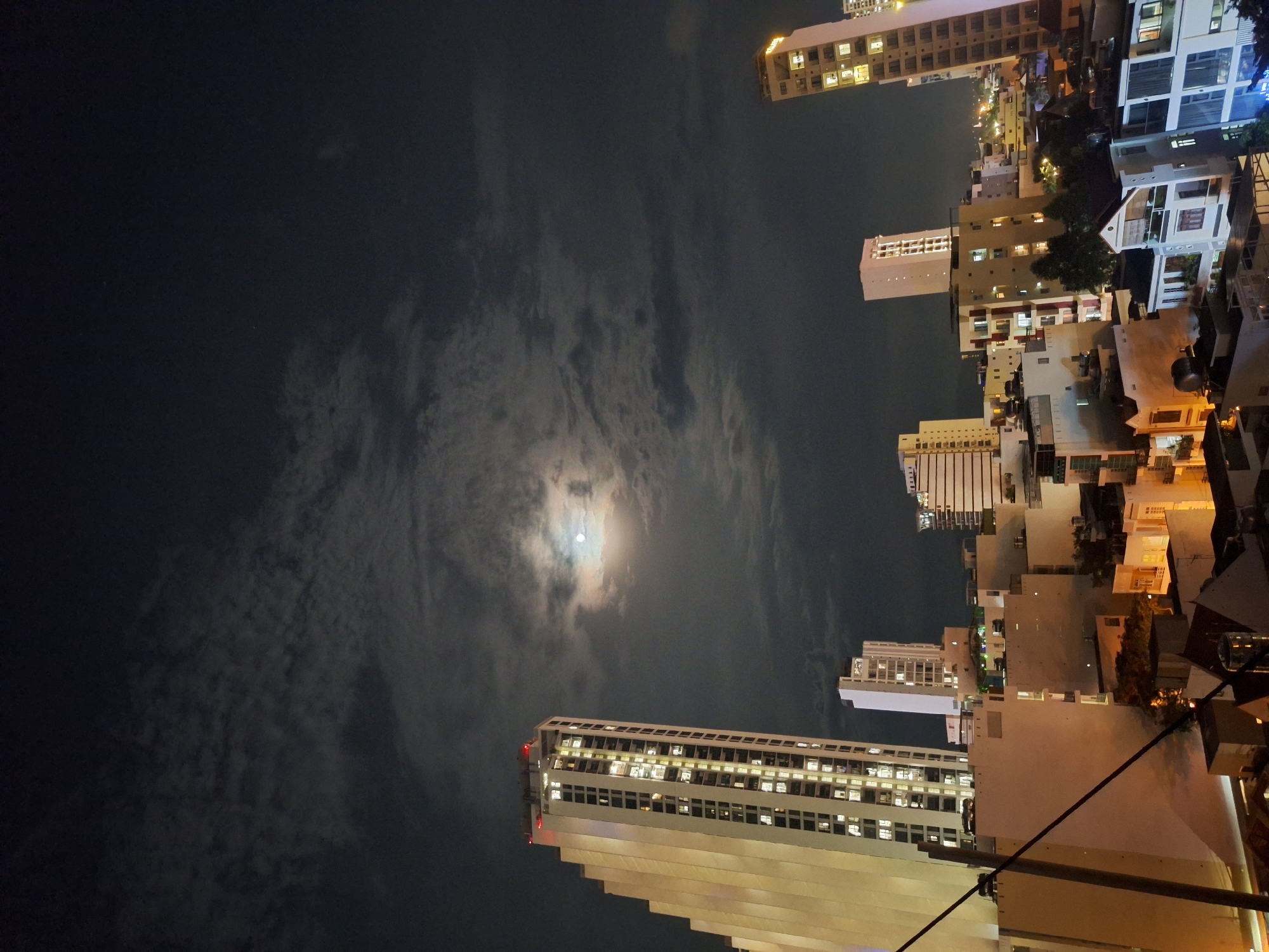 The image shows a nighttime cityscape in Nha Trang, Vietnam, under a cloudy sky with a visible moon. The scene features several high-rise buildings, including a prominent white skyscraper on the left with illuminated windows. In the distance, more buildings and lights of the city are visible. The overall atmosphere is urban and nocturnal.