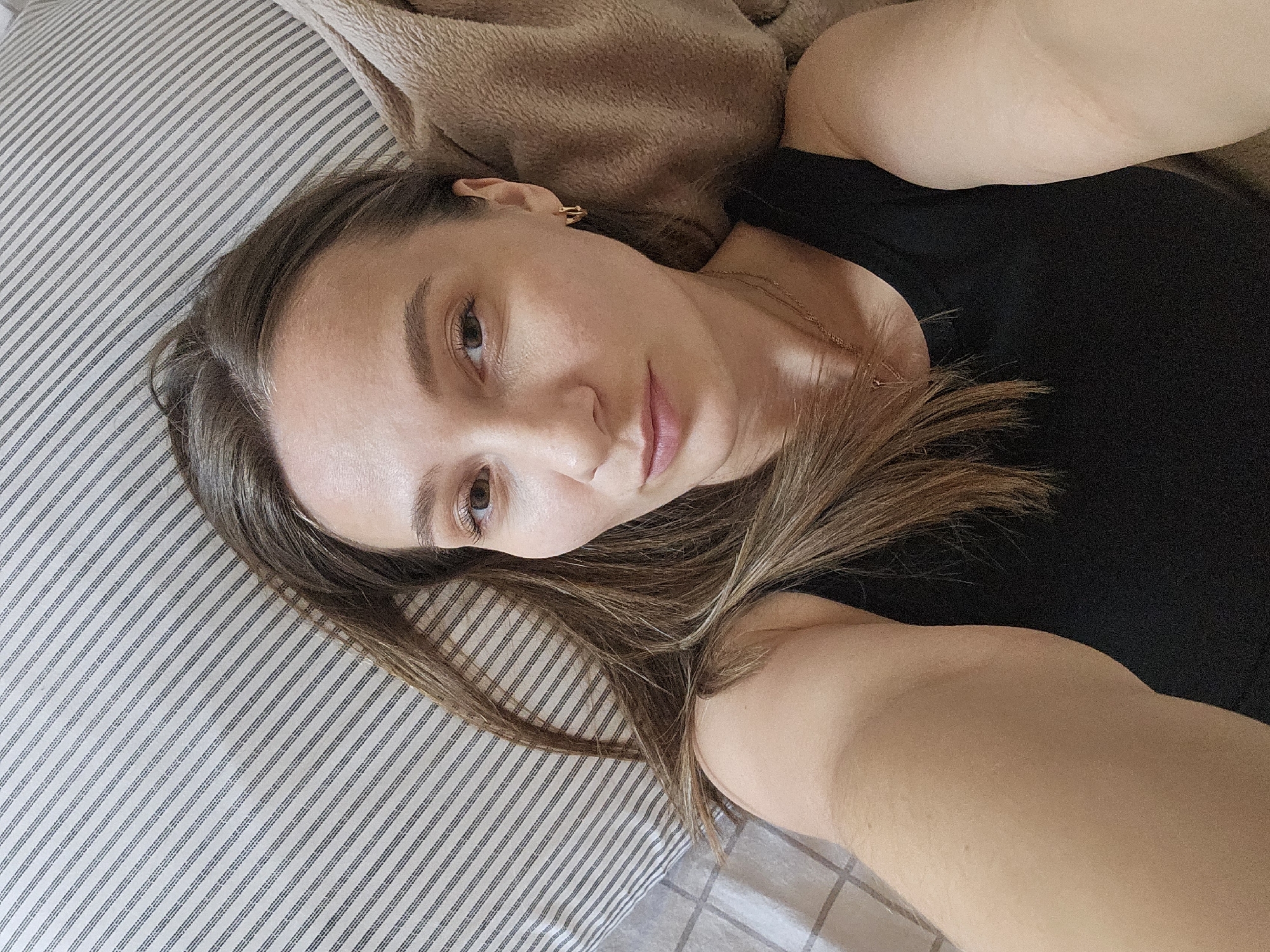 A woman is lying on a striped pillow with a tan blanket near her head. She is wearing a black top and a gold necklace, and she has gold earrings. Her hair is brown with lighter highlights. The location is not discernable from the image.