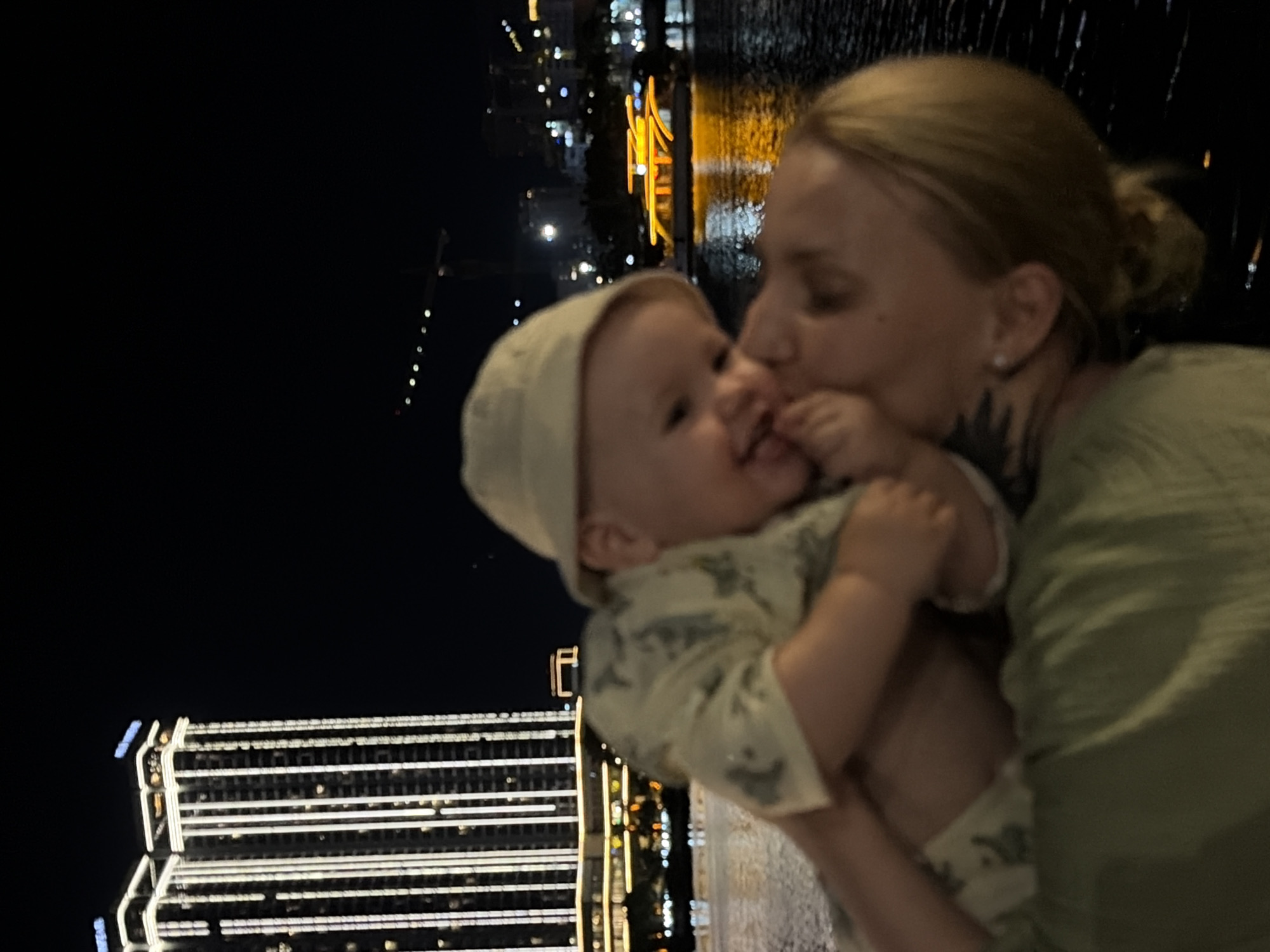 The image portrays an intimate moment between a mother and her baby at night in Đà Nẵng, Vietnam. The mother, with blonde hair pulled back, occupies the right side of the frame, wearing a light green top. She is captured in the act of tenderly kissing her baby on the cheek or mouth, with a tattoo visible on her neck/shoulder area.
The baby, positioned centrally and to the left, wears a light-colored bonnet or hat and a patterned romper. The baby's face is slightly out of focus but appears to be looking towards the mother, with small hands near the face.
The background reveals a bustling city at night. To the left, a modern, illuminated building with multiple rows of white lights on its exterior stands out. In the mid-ground and extending to the right, a body of water, likely a river or bay, reflects the vibrant city lights in streaks of orange, yellow, and white. Numerous other distant lights from buildings and possibly streetlights are visible, creating a luminous urban backdrop. The setting strongly suggests an evening outing in a lively city. The expressions and posture convey a feeling of warmth and affection between the mother and child. There is no visible text in the image.