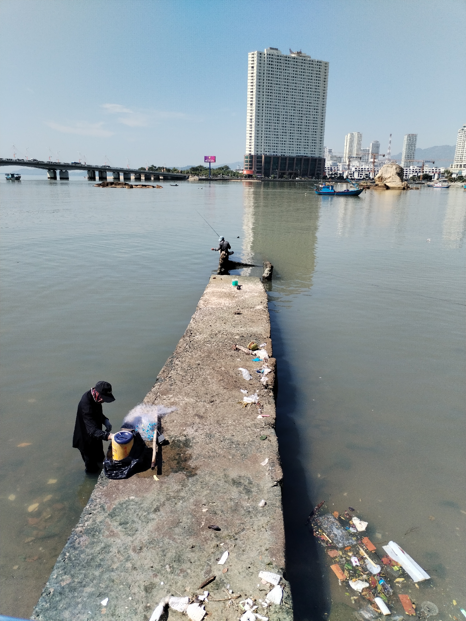 The image depicts a waterfront scene in Bac Nha Trang Ward, Vietnam. A concrete pier juts into a body of water, with a man standing in the water at the pier's edge. He is wearing dark clothing, including a jacket and cap, and appears to be using a net or similar equipment. A yellow bucket sits beside him, along with other equipment. Further along the pier, another person is fishing. The water around the pier is polluted with trash. In the background, a bridge spans the water, and a tall, white building dominates the skyline along with other buildings. The sky is clear and blue.