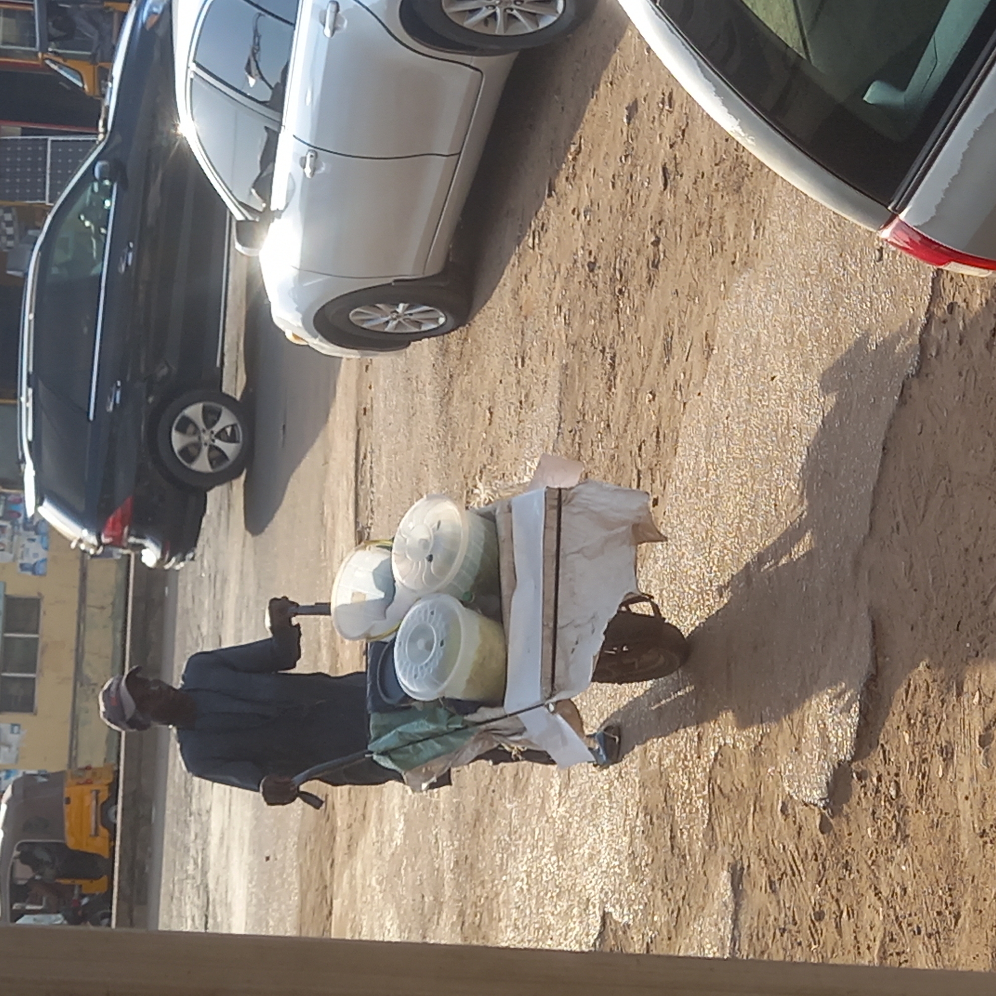 In Sardauna, Nigeria, a man wearing a dark outfit and cap pushes a small, two-wheeled cart. The cart carries several containers with white lids, partially covered by a white sheet or cloth. The environment appears to be a street with at least two cars parked nearby: a dark SUV and a light-colored sedan. The ground is a mix of concrete and dirt. In the background, a yellow auto-rickshaw is visible, and there are buildings lining the street.