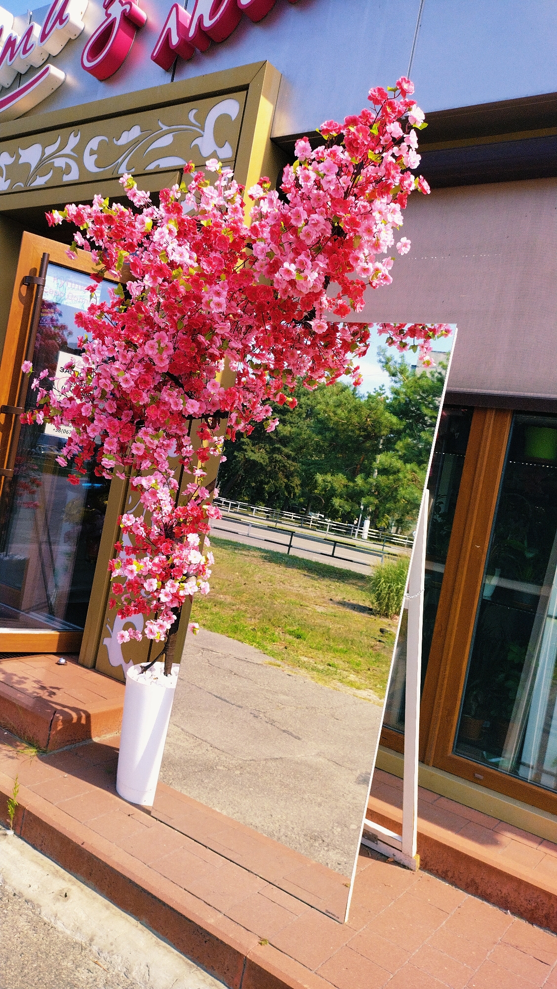 A fake flowering tree with pink and red blossoms is positioned in front of a building. The tree is in a white cylindrical planter filled with white stones. The building has a decorative gold trim with white floral designs and a sign with pink lettering that reads "МАГІЯ ЗЕЛЯ". To the right of the tree, a tall rectangular mirror leans against the building, reflecting the sky, trees, and a white fence. The scene is set outdoors, with a concrete sidewalk and a grassy area visible. The building appears to be a commercial establishment. The location is specified as Horishni Plavni, Ukraine.