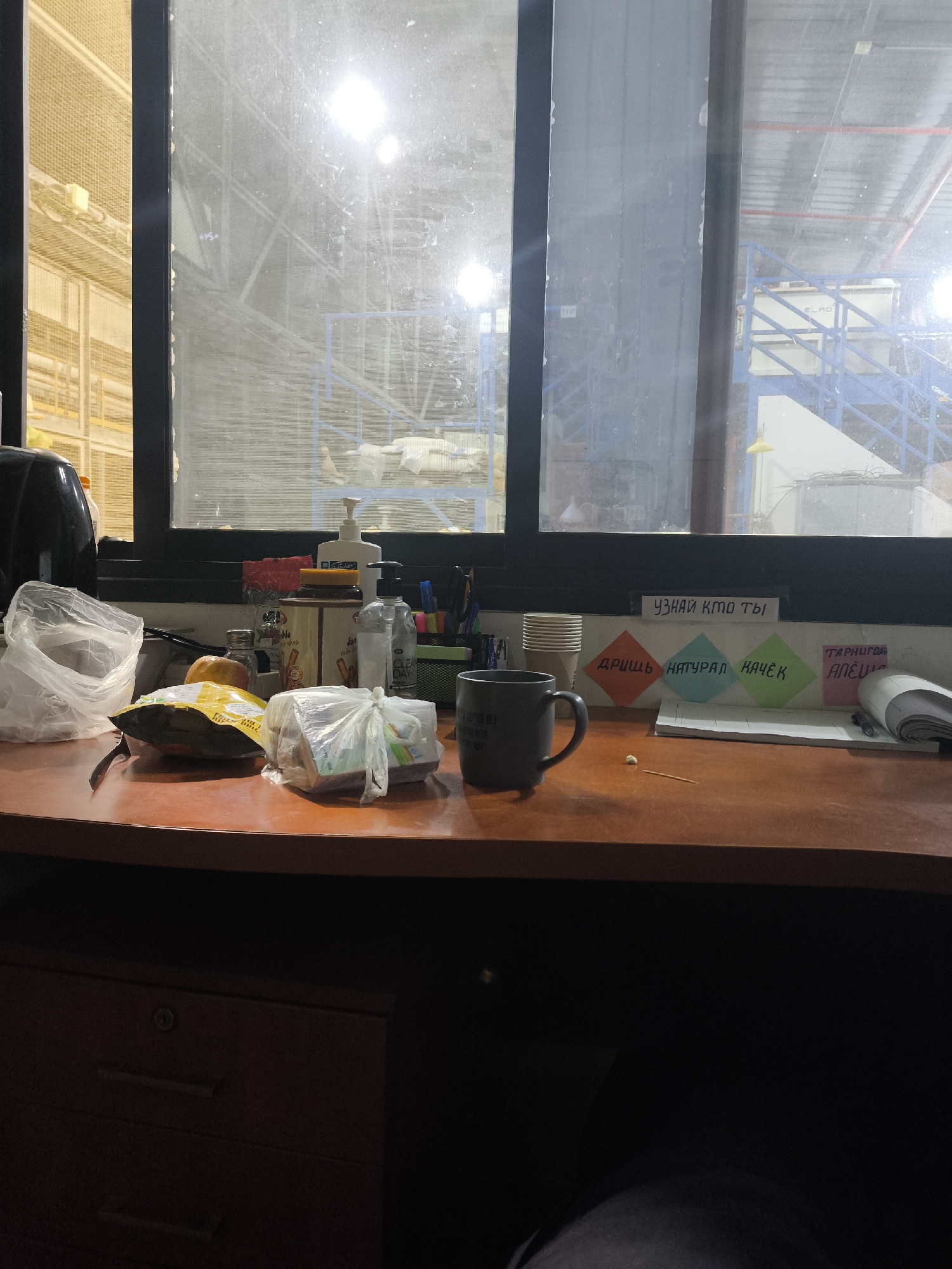The image depicts a desk inside a dimly lit room with a large window. The window offers a view of what appears to be a warehouse or industrial facility. Outside, there are rows of yellow shelving units and blue metal structures, possibly part of a storage or manufacturing complex. The window panes are grimy and reflect the interior lights and some of the exterior environment.
On the desk, several objects are visible: a black mug with a handle, a stack of white paper cups, a notebook with a rolled-up section, several colorful sticky notes, and a small toothpick with a white speck on it. There are also various items in plastic bags, including what looks like food packaging and personal care items like a bottle of hand sanitizer labeled "CLEAR DAY". A yellow apple and an orange are also present on the desk. A black electrical appliance, possibly a kettle, is partially visible on the left side of the desk. A dark wooden drawer unit is beneath the desk.
The sticky notes on the wall above the desk have Russian text. One note reads "УЗНАЙ КТО ТЫ" which translates to "Find out who you are". Below this, three diamond-shaped notes display words like "ДРИЩЬ", "НАТУРАЛ", and "КАЧЁК", which are informal Russian terms related to body types. A fourth note, pink in color, has text that is difficult to decipher but appears to be in Cyrillic script.
The overall impression is a workspace that is somewhat cluttered and personal, set against the backdrop of a large industrial space, suggesting it could be an office or break area within a facility in Dimona, Israel. The lighting is artificial and casts shadows, contributing to a slightly somber atmosphere. There are no visible people in the image.
