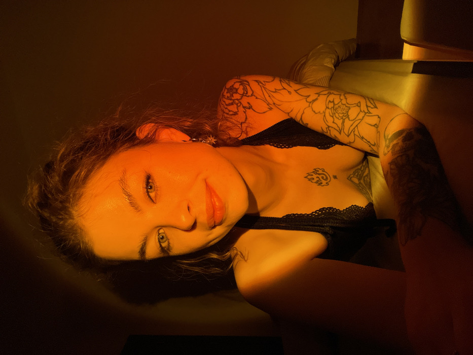 A woman lies with her head tilted on a surface, bathed in warm, orange light. She wears a black, lacy top. Tattoos adorn her upper arms and chest. She gazes directly at the viewer with a slight smile, her eyes appearing green. The lighting creates dramatic shadows, adding depth to the image. Part of a bed or sofa is visible in the background.