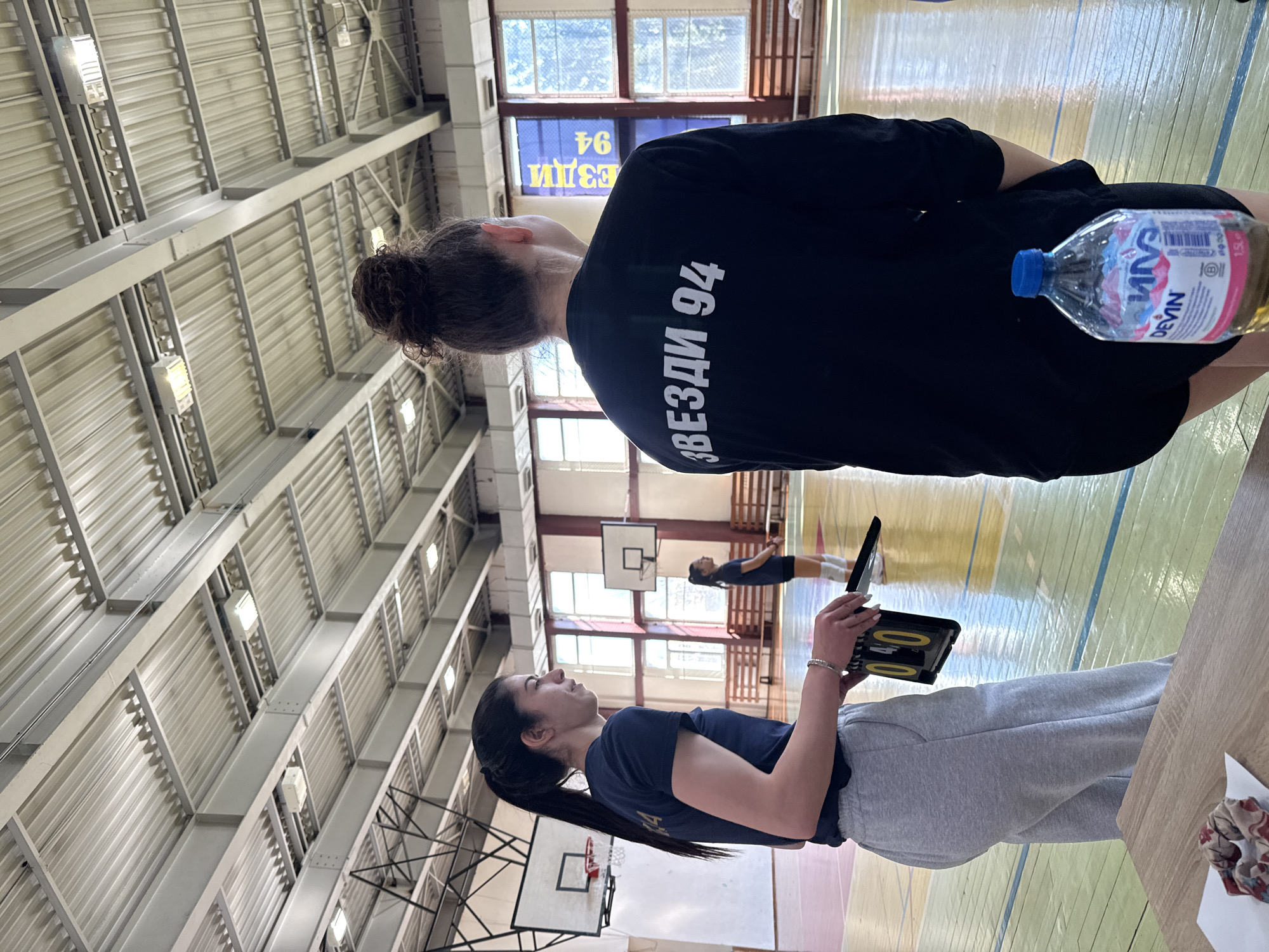The image shows the interior of a gymnasium in Sofia, Bulgaria. Two women are prominently featured. One woman, seen from the back, wears a black shirt with the text "ЗВЕЗДИ 94" and the number "94" printed in white. A bottle of "Devin" water is placed near her. The other woman is holding a scoreboard displaying "040." She wears a dark blue shirt and gray sweatpants. A basketball court with hoops is visible, with a person playing basketball in the background. The gymnasium has a metal ceiling structure with light fixtures. Windows are visible near the top of the frame.