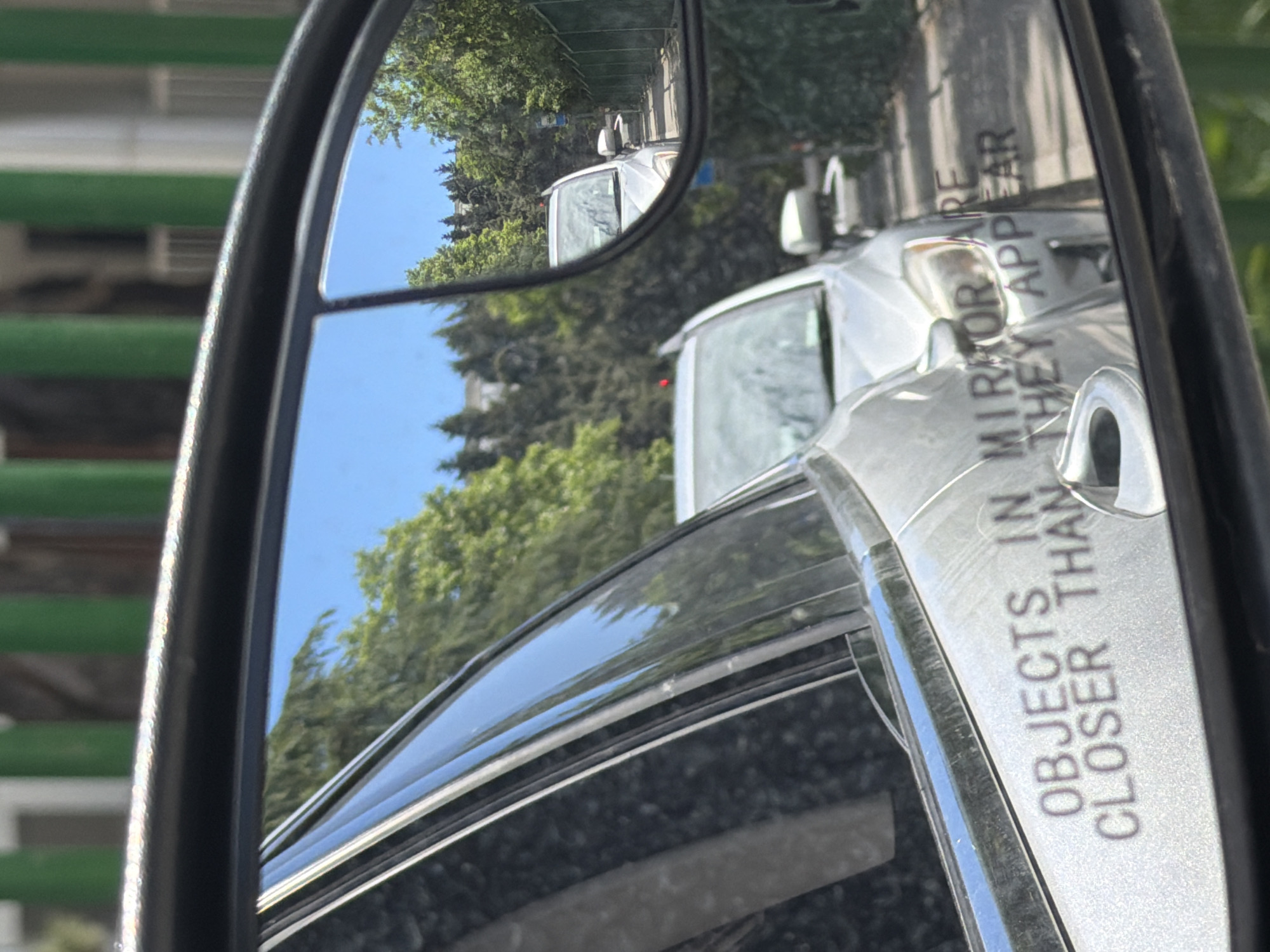 The image shows a side view mirror of a car. The mirror reflects the image of a white van on a street lined with trees. The mirror housing has the warning "OBJECTS IN MIRROR ARE CLOSER THAN THEY APPEAR". The background outside the car shows a green and white building. The location is in Tbilisi, Georgia.