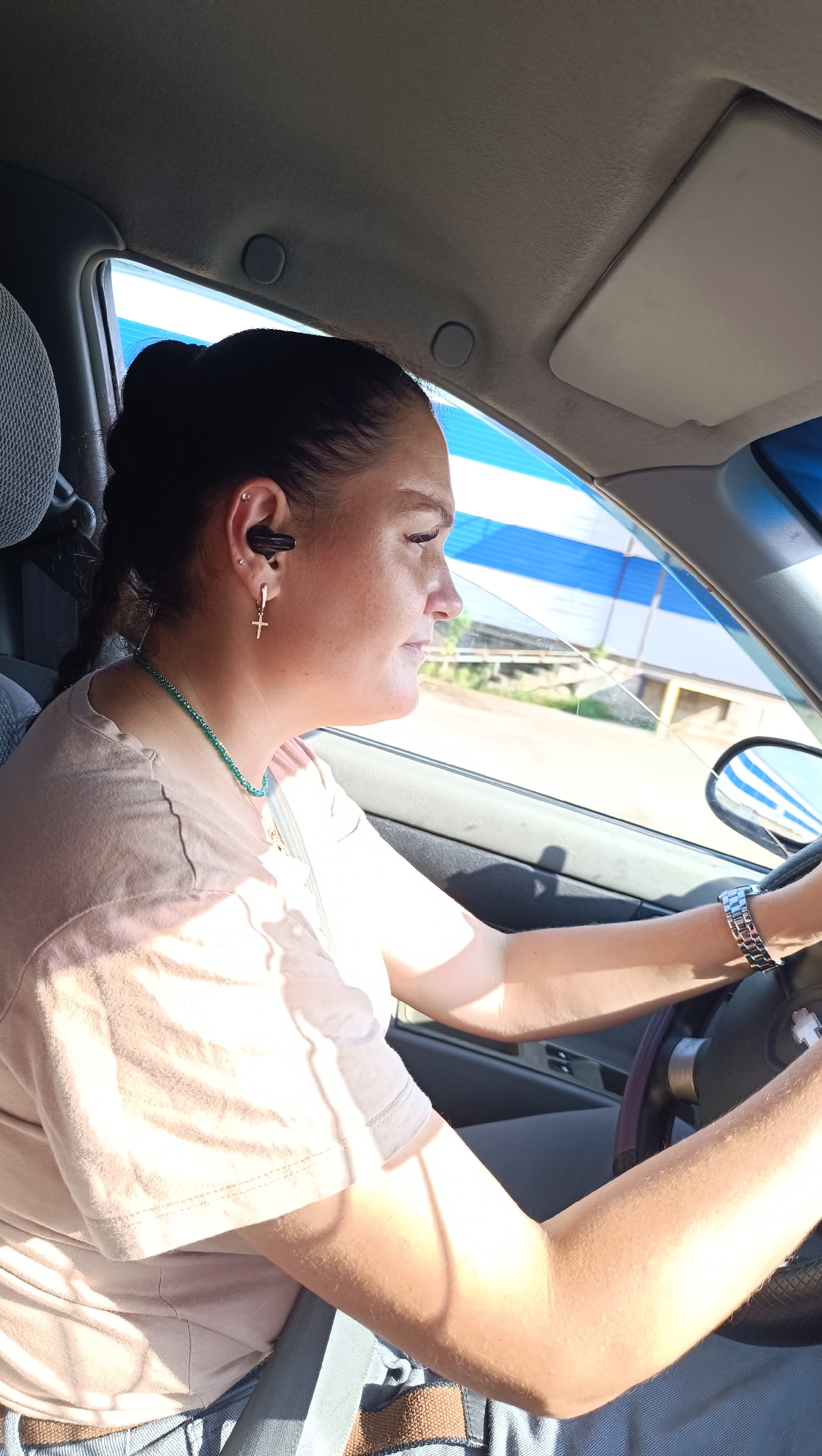 A young woman is driving a car on a sunny day in Chaikovsky, Russia. She has dark hair pulled back in a bun and a braid, and is wearing a light pink t-shirt and blue jeans. She has a silver stud in her left ear and a gold cross earring. She is also wearing a turquoise beaded necklace and a silver watch on her left wrist. The interior of the car is visible, including the steering wheel, dashboard, and a portion of the passenger seat. The woman is looking forward, presumably at the road ahead. The background outside the car shows a blue and white striped structure and some trees, suggesting an outdoor environment.