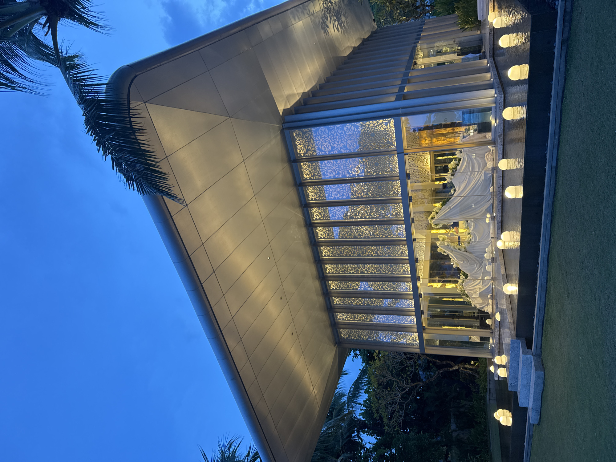 Captured at dusk, the image showcases a modern architectural structure located in Kuta Selatan, Indonesia. The building features a sloping roof with integrated lighting and a glass facade adorned with decorative, patterned screens. Inside, preparations appear to be underway, possibly for a ceremony, evidenced by draped white fabric and floral arrangements. A narrow water feature runs along the building's exterior, punctuated by evenly spaced, illuminated globes. To the side of the building, well-manicured grass provides a contrasting texture. Palm trees are visible in the foreground and background, characteristic of the tropical environment. The sky transitions from light to deep blue, indicating the fading daylight.