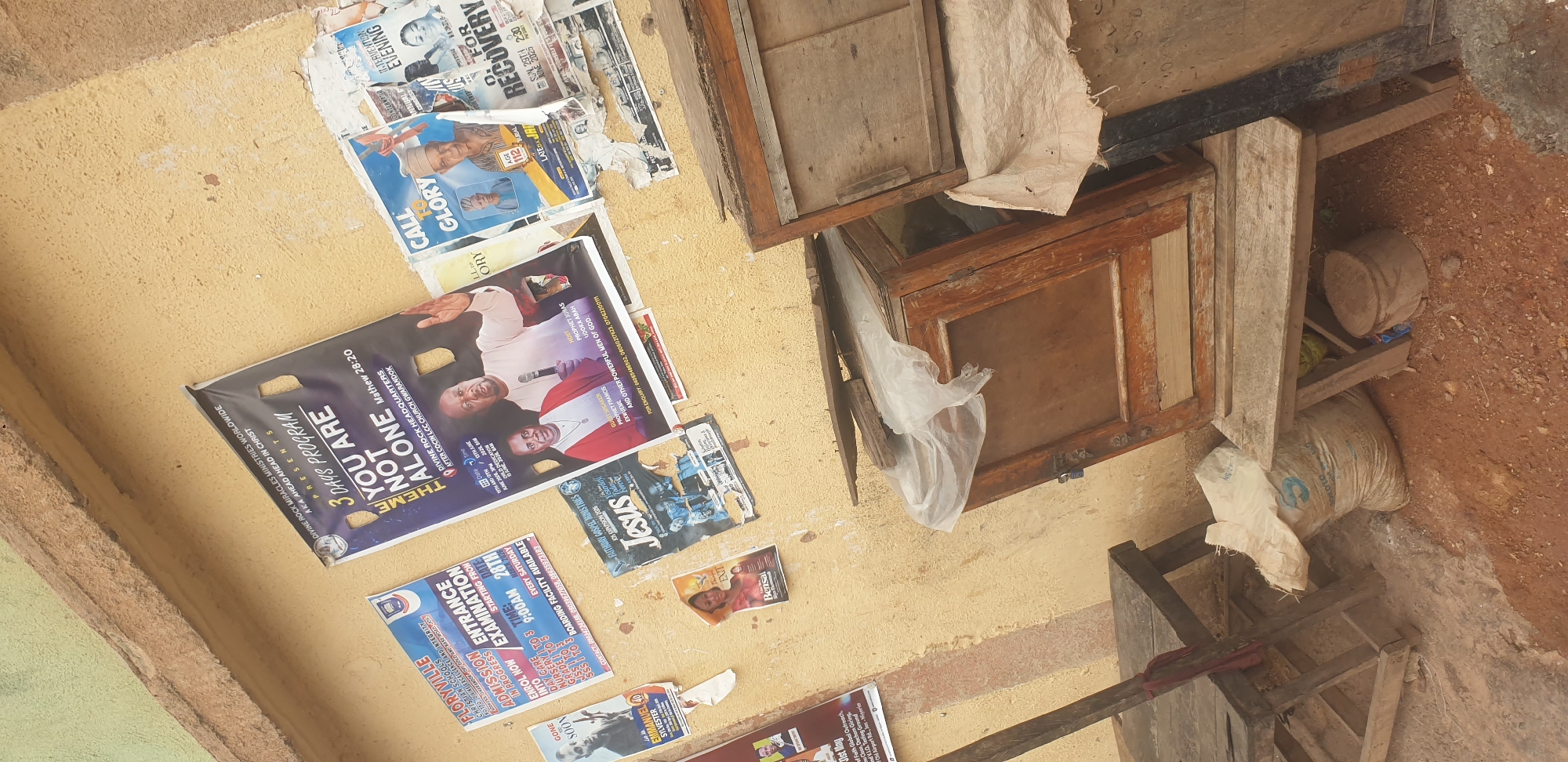 This is a photograph taken outdoors in Barakin Lerawi, Nigeria, focusing on a textured, pale yellow wall covered in various posters and flyers. In the foreground, two worn, wooden benches are visible, one on the left and another on the right. Between the benches, a wooden cabinet with glass doors is placed on top of another wooden structure. A white plastic bag is draped over the cabinet and a larger sack, likely containing goods, rests on the ground near the left bench. The ground appears to be dirt and gravel.
The wall displays a collage of printed materials. Prominently featured is a large poster with the text "3 DAYS PROGRAM YOU ARE NOT ALONE" and a Bible verse reference, Matthew 28:20. This poster also depicts two individuals, likely religious figures. To its left, a flyer for "FLORNYVILLE CHRISTIAN SCHOOLS" announces "ENTRANCE EXAMINATION IN PROGRESS." Other smaller posters and flyers are scattered across the wall, some partially torn or obscured. One poster, with a picture of a man, has the text "GONE SOON" and "EMMANUEL." Another flyer shows the word "Jesus" prominently. A flyer with "CALL TO GLORY" and "AGE 112" is also visible, as is a poster advertising an "INTERVENTION EVENING." The overall impression is of a community space where events and announcements are shared through public postings.