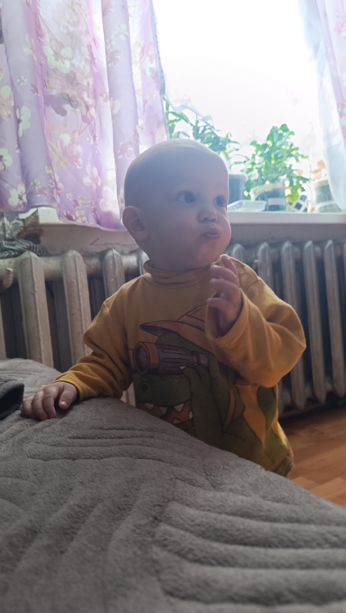 A young child, appearing to be between one and two years old, is standing and leaning on a textured grey blanket. The child has light skin, a bald head, and is wearing a yellow long-sleeved shirt with a dinosaur graphic. The child's face is turned to the right, with cheeks puffed out and lips pursed. One hand is resting on the blanket, while the other is raised slightly. In the background, a white radiator runs along the wall. Behind the radiator, sheer purple and white curtains with a floral pattern hang in front of a window, through which plants are visible. The scene is indoors, suggesting a residential setting, potentially in Karsha, Russia, given the architectural style of the radiator and curtains which are common in Russian apartments.
