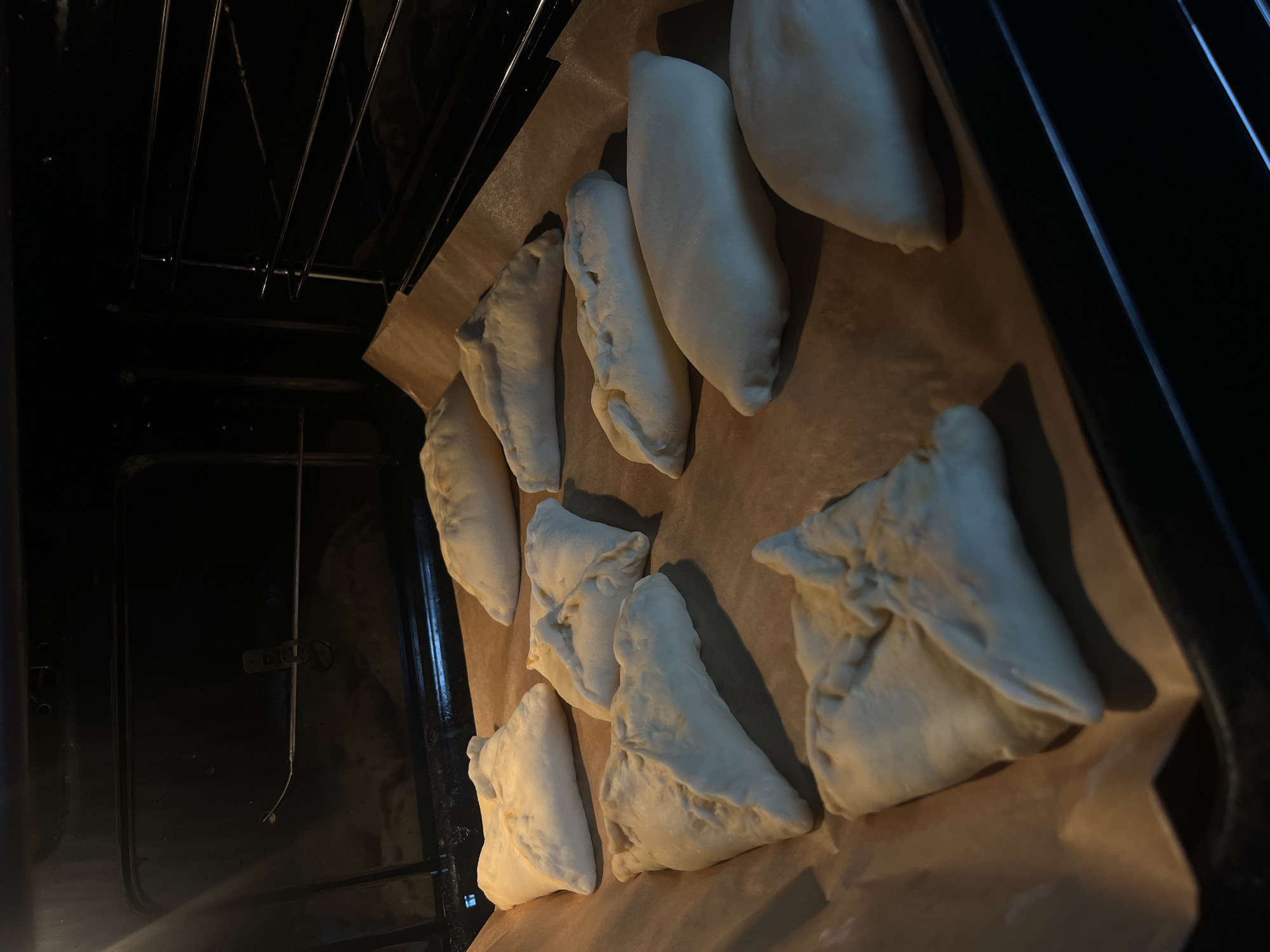 The image shows a baking sheet lined with parchment paper inside a dark oven. There are several uncooked pastries on the baking sheet. The pastries are arranged in rows and are various shapes, some elongated and others more triangular or square. The oven interior is dimly lit, and the oven rack is visible. The location, Shushary, Russia, is not discernible from the image.