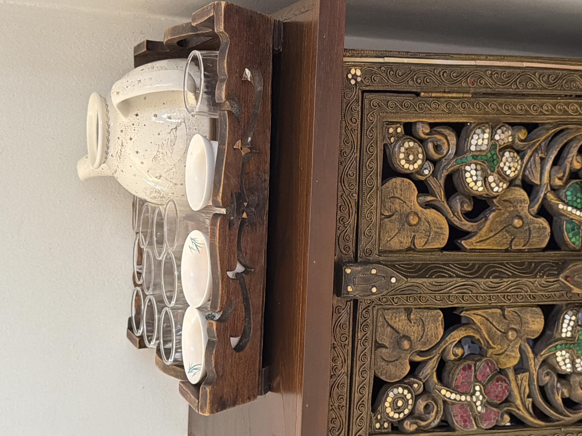 In Ko Samui District, Thailand, a wooden tray filled with a tea set rests on a dark wooden cabinet. The tray holds a white ceramic teapot with a speckled pattern and a handle, along with several clear glass cups. Two small white bowls with green bamboo designs are also present in the tray. The cabinet below is ornately carved with floral and swirling motifs, featuring inlaid colorful stones and metal accents. The background consists of a plain, light-colored wall.