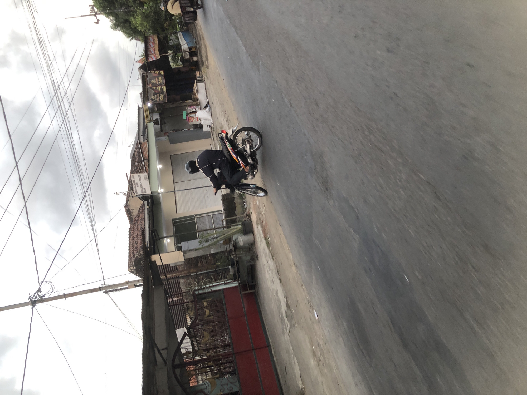 In the city of Kesambi, Indonesia, a person wearing a black helmet and jacket rides a motorcycle on a paved road. The motorcycle is black with colorful decals on its side and has a silver exhaust pipe. The rider is looking forward. To the left of the road, there is a building with a red gate and a tiled roof. Various shops with awnings are visible in the background, one displaying advertisements with the numbers "20/10" and "2,000." A power pole with numerous overhead wires stands prominently on the left side of the frame. The sky is overcast with a mix of clouds. Another person on a motorcycle with a side trailer is seen in the distance on the right side of the road. The overall environment appears to be a commercial or residential street.