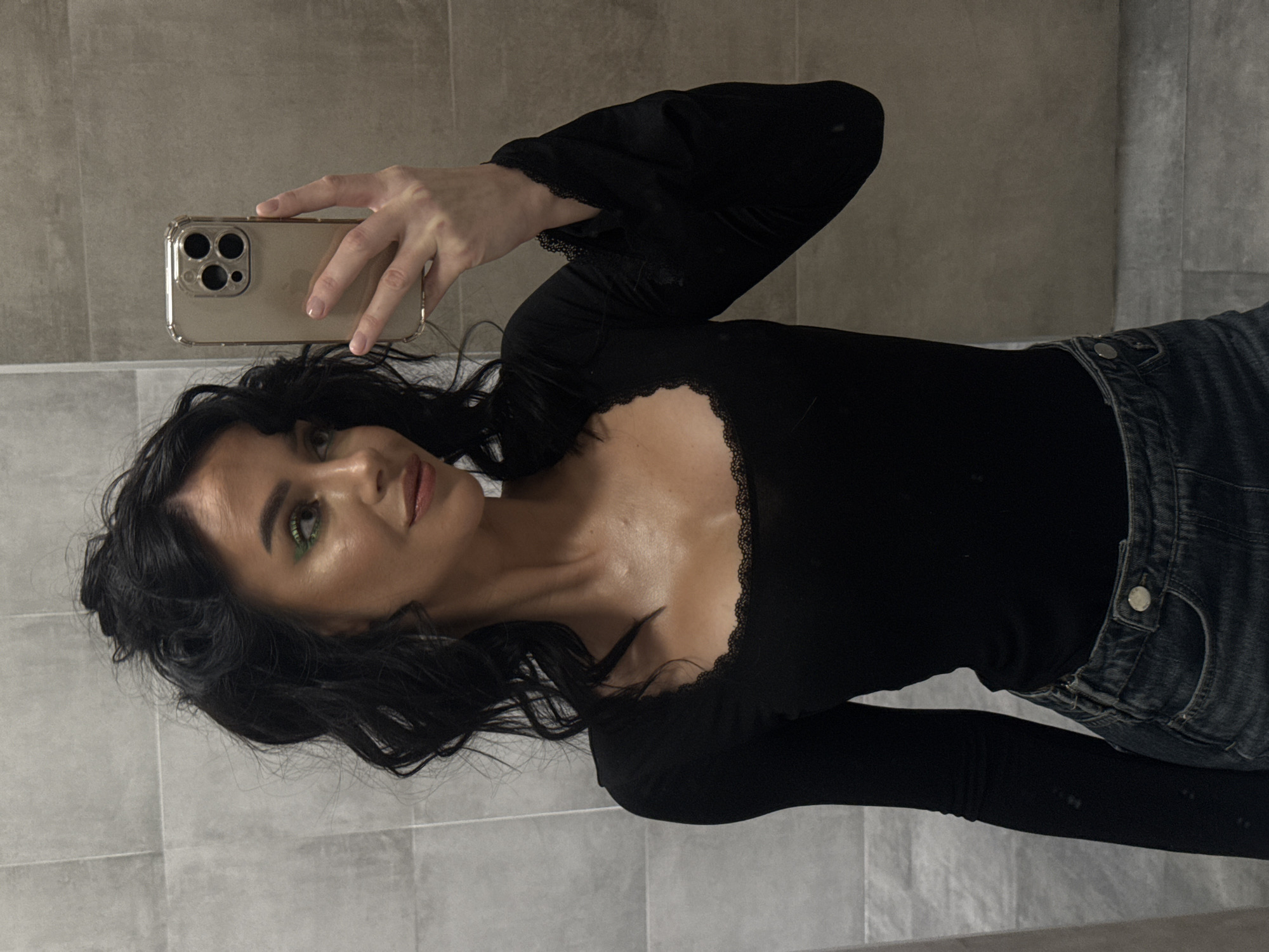 The image displays a mirror selfie of a woman, oriented 90 degrees clockwise. The woman has dark, wavy hair and is looking towards her reflection with a subtle smile. She wears green eyeshadow and a nude lip color. Her attire consists of a fitted black long-sleeved top featuring a sweetheart neckline and cuffs, both adorned with black lace trim. Dark wash denim jeans are visible on her lower body.
She is holding a gold-toned iPhone, likely a Pro model due to the prominent triple camera module, enclosed in a clear protective case. The word "iPhone" is visible on the back of the device. The background shows light gray rectangular tiles with darker grout lines, characteristic of an indoor setting such as a bathroom or a tiled wall. The lighting appears even and diffused, typical of interior artificial light. The image lacks specific details regarding the time of day or weather. The provided context indicates the location is Krasnoyarsk, Russia.