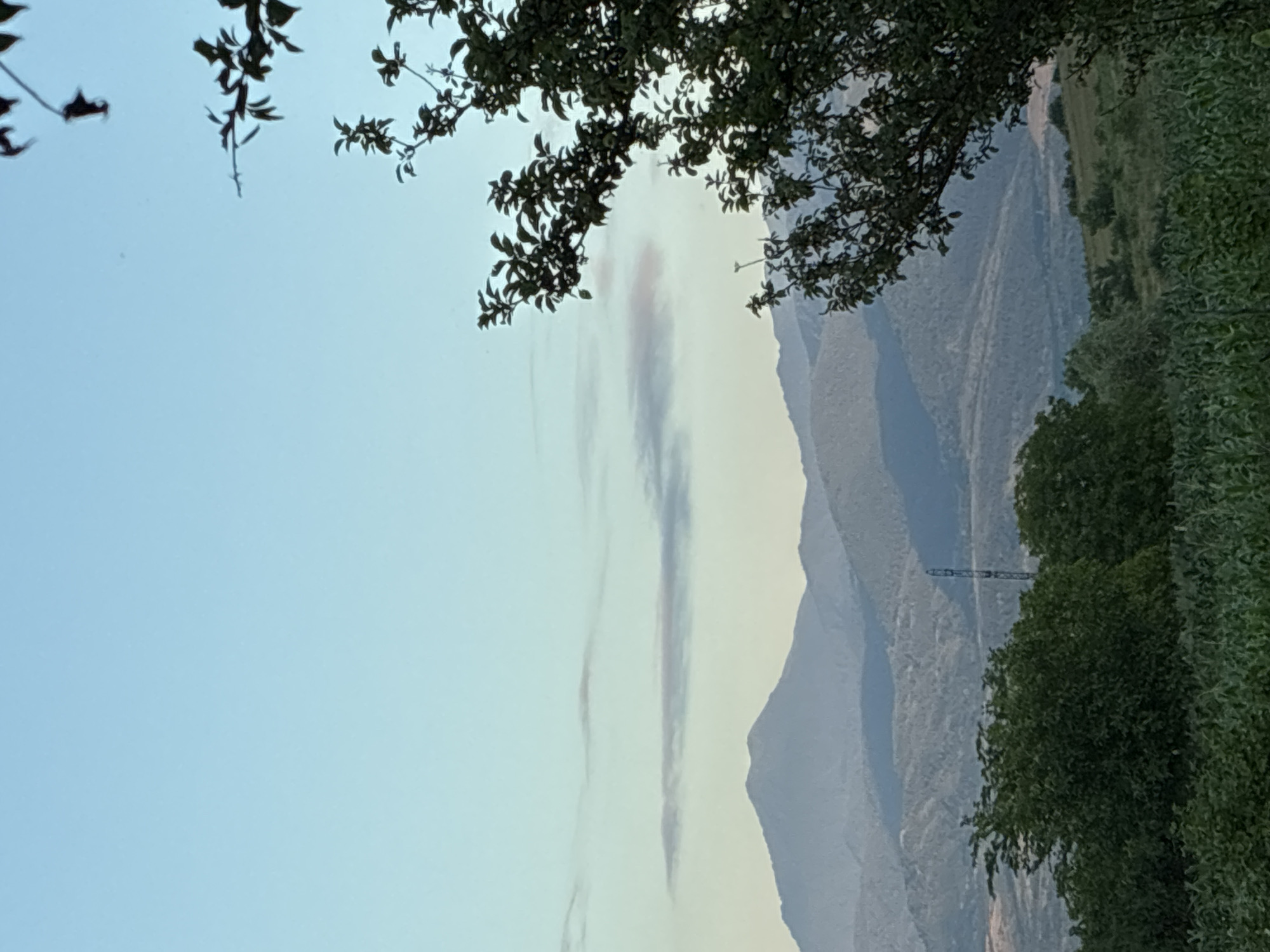 This media file captures a serene natural landscape, likely in Surami, Georgia. The scene is dominated by lush green trees framing the foreground, with their leaves silhouetted against a pale blue sky. In the distance, a series of layered mountains recede into the horizon, their slopes softened by the atmospheric haze. Wispy, elongated clouds streak vertically across the sky, adding a dynamic element to the otherwise still vista. A tall, slender tower, possibly a telecommunications mast or a part of a distant structure, is visible nestled within the mountain range. The overall impression is one of tranquility and natural beauty, with the golden hour light hinting at either dawn or dusk. There is no visible text in the image.