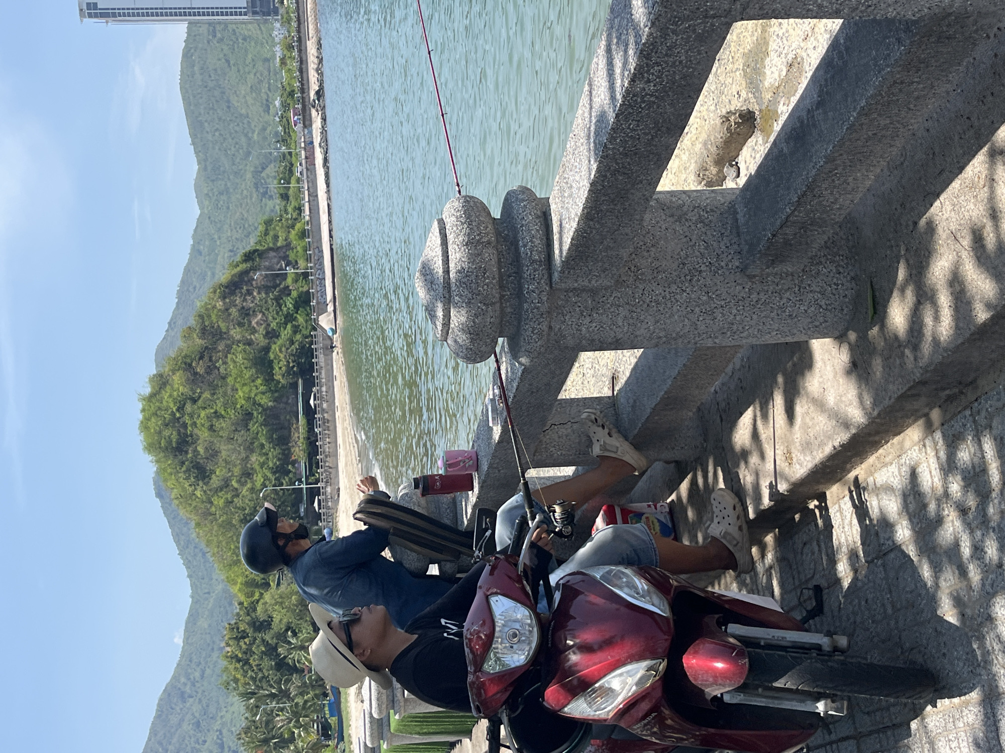 The image captures a sunlit coastal scene in Bac Nha Trang Ward, Vietnam, featuring two individuals fishing from a granite promenade. In the immediate foreground, a red Honda scooter is parked, facing away from the water. Seated on the scooter is a person wearing a light fedora, sunglasses, a dark long-sleeved shirt, and light-colored shorts; their left foot, clad in a white Croc-style sandal, is visible. This individual holds a fishing rod extending over the granite railing into the water. Behind and to the right, a second person, wearing a dark helmet and a dark long-sleeved shirt, leans against the railing, also appearing to fish. A white Croc-style sandal is visible on their left foot. Two beverage containers, one red and one pink, and a striped bag are positioned on or leaning against the granite railing near the second individual.
The promenade railing is constructed of speckled grey granite, featuring ornamental balusters. Beyond the railing, the calm, light blue-green water of a bay or inlet is visible. A narrow strip of light-colored sand and rocks forms a beach at the water's edge. Further inland, a paved road with discernible vehicles and streetlights runs parallel to the coastline. Lush, green, forested mountains dominate the background, extending across the horizon. On the far right, a tall, modern high-rise building is visible. The sky is clear blue. Strong, distinct shadows from the railing and potential overhead foliage are cast upon the promenade, indicating bright, midday sunlight. Small blue boats are visible on the distant sandy shore to the left.