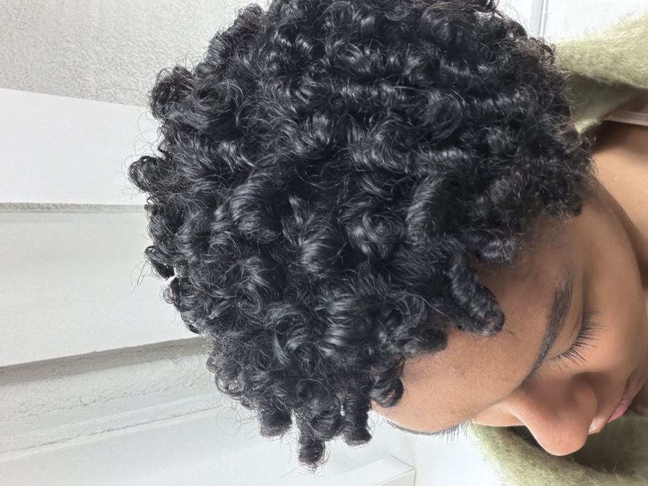 The image captures a high-angle, close-up perspective of the left side of an individual's head and upper torso. The subject has dark, tightly coiled, well-defined curly hair, appearing hydrated or styled. Their skin tone is medium-dark. The left eye is partially visible, appearing closed or directed downwards, with distinct eyelashes and an eyebrow. The bridge of the nose is also visible. The individual wears a textured, olive-green outer garment, possibly knitted, over a lighter, possibly tan or nude, inner layer. The background consists of plain, light-colored vertical surfaces, indicative of an indoor environment with architectural features. No visual information within the image provides specific geographic markers or details to identify a location in Paris, France, or any other city.