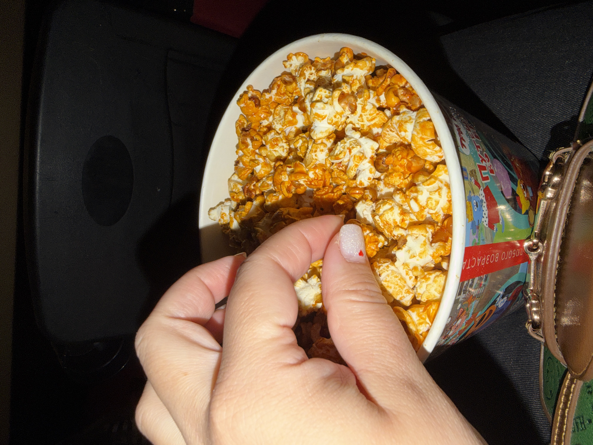 The image shows a person reaching into a container of caramel popcorn. The container is white and decorated with cartoon characters. The person's hand has a nail with a red heart design. The background is dark, with a black object on the left and part of a purse on the right. The location is unknown.