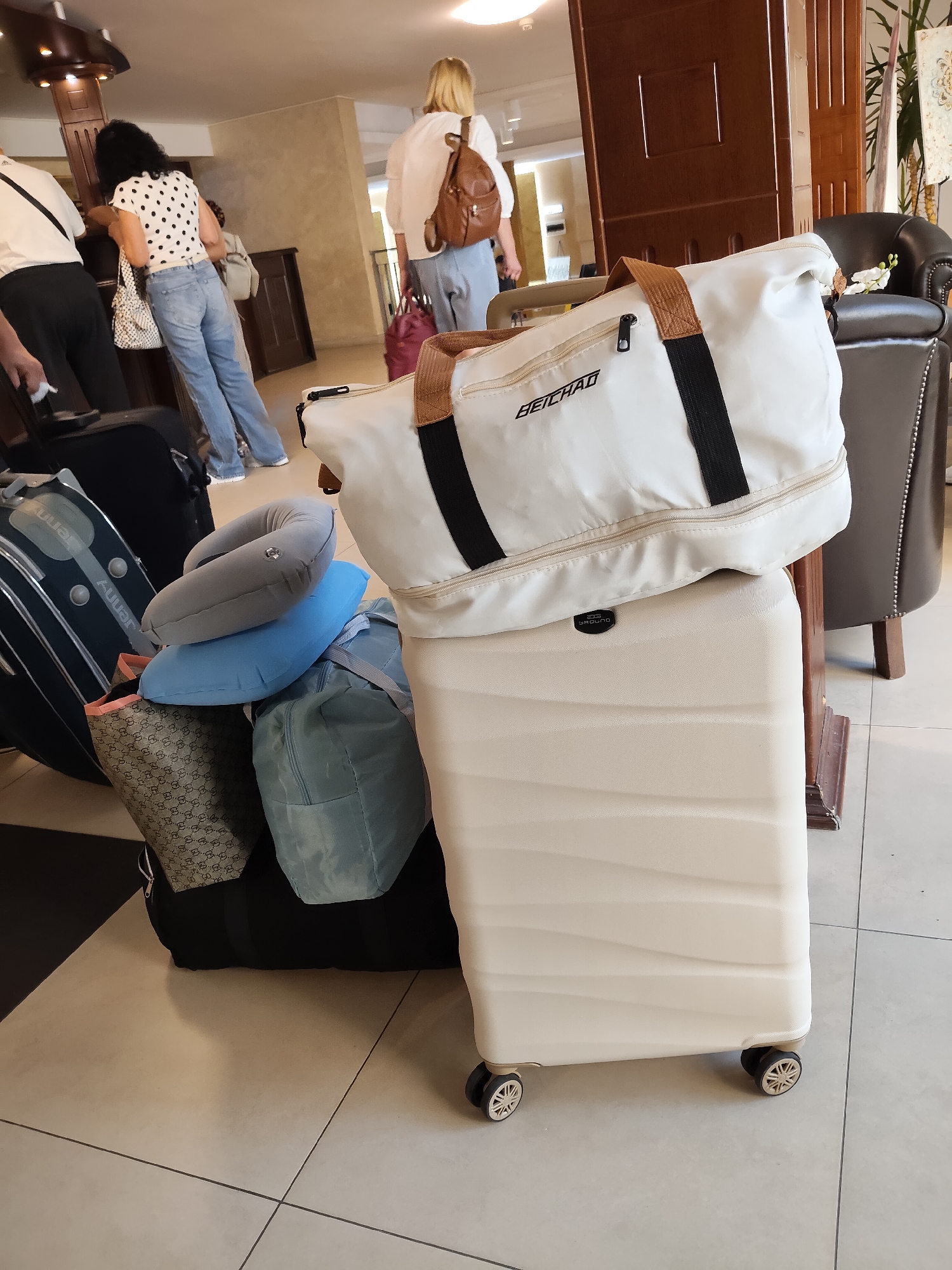 A hotel lobby scene features several people and a large collection of luggage. In the foreground, a light-colored hard-shell suitcase stands upright, topped with a white duffel bag with black and brown accents. Next to it, a stack of bags includes a patterned tote, a blue pillow, a lighter blue pillow, and a teal duffel bag. To the left, a black suitcase with silver accents is partially visible. In the background, two women are at a reception desk. One woman, wearing a white polka-dot shirt and jeans, is leaning on the counter. The other woman, wearing a light-colored shirt and jeans, has a brown backpack and is holding a pink bag. A man in a white shirt and black pants stands to the far left, holding a black bag. The environment appears to be a hotel lobby with tiled flooring, wooden furniture, and a leather armchair.