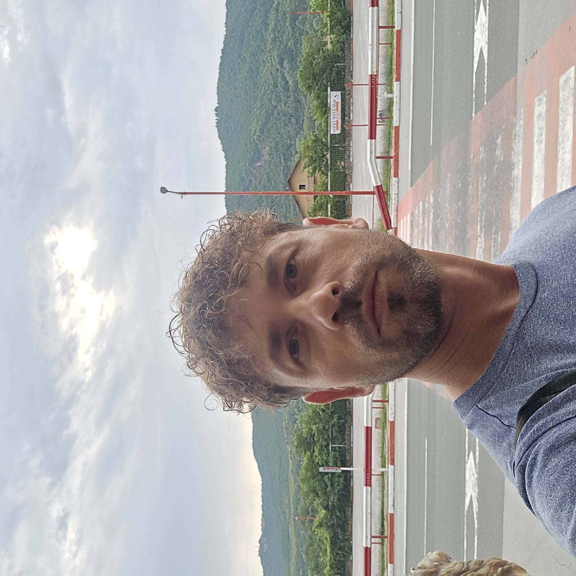 A man with curly brown hair and a beard stands in the foreground, taking a selfie. He is wearing a blue t-shirt. Behind him, a road with white and red barriers is visible, with a pedestrian crossing marked by red and white stripes. In the background, a lush green mountain looms under a cloudy sky with the sun partially visible through the clouds. A street lamp with a red pole is also present in the background. A building with a brown roof and green trees surround it is visible on the slope of the mountain. A sign with text in an unfamiliar alphabet is mounted on a fence. The scene appears to be in Zlatna Panega, Bulgaria.
