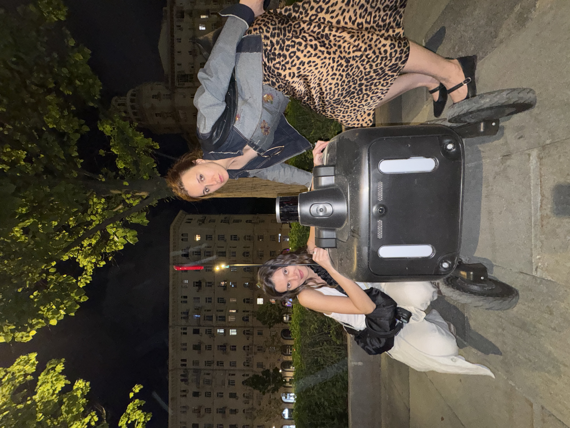 Two women pose behind a food delivery robot on a city street at night. One woman, with reddish-brown hair, wears a denim jacket with leather sleeves and a leopard-print dress. The other woman, with dark hair, wears a white top and white pants, with a black bag on her shoulder. The delivery robot is black with two wheels, and it has a camera on top. The background includes a building with many windows. Based on the architecture, lighting, and other visual elements, the location is likely Moscow, Russia.