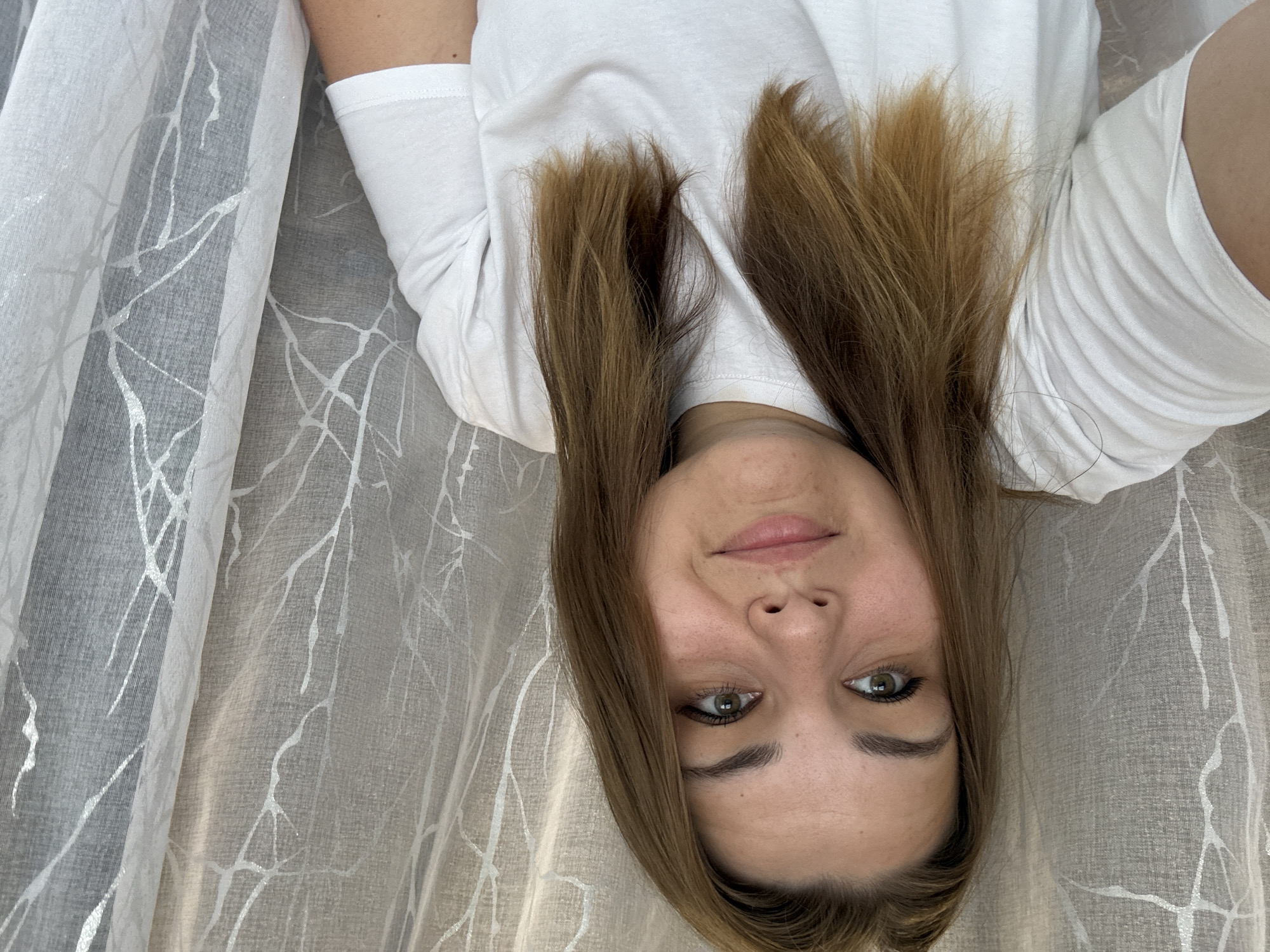 A young woman is lying down, taking a selfie. She has light brown hair and green eyes. She is wearing a white T-shirt. She is lying on a light grey fabric with a white branch pattern. The location is not specified, it is unknown if this photo was taken in Chaikovsky, Russia.