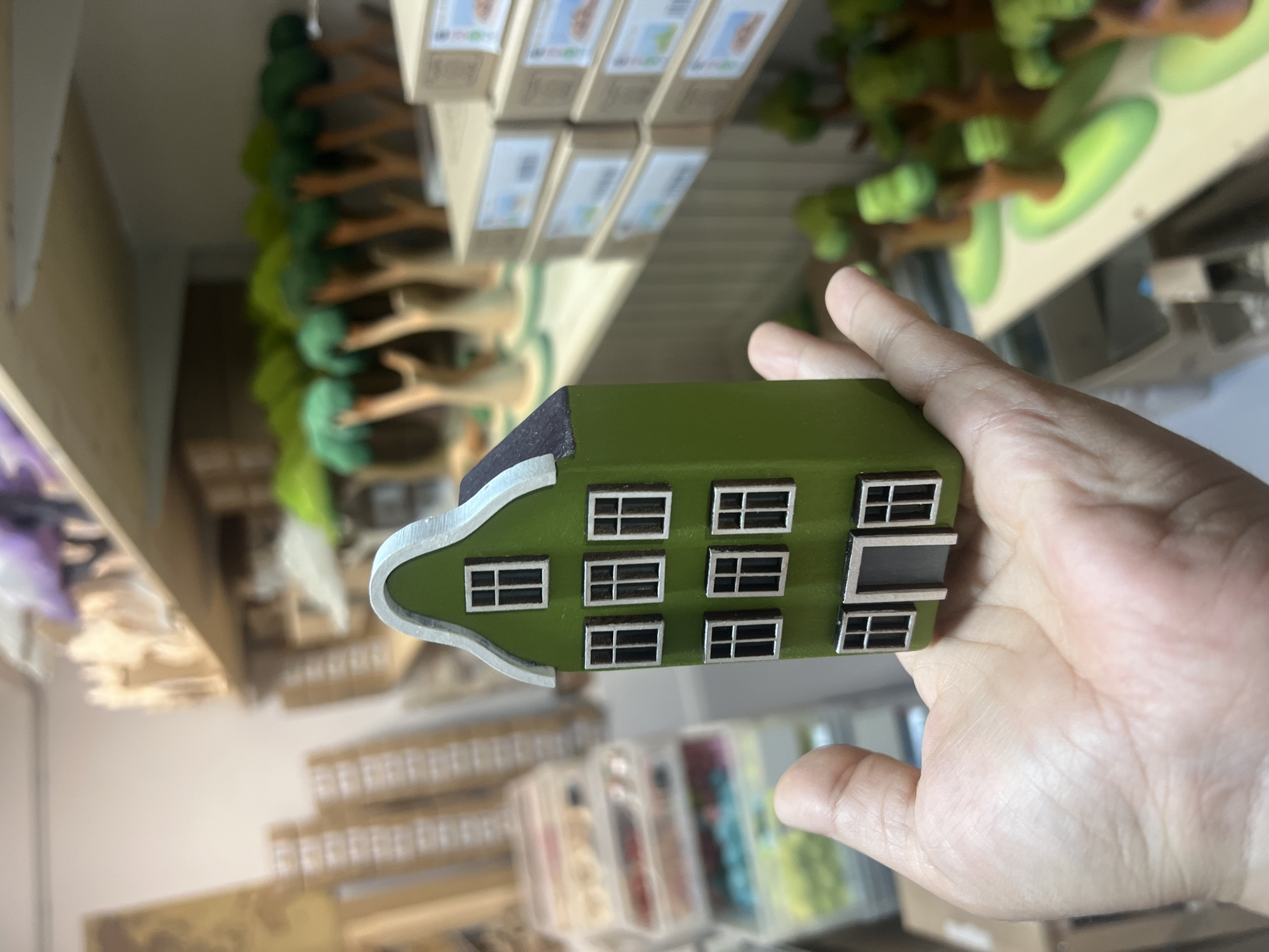 A hand is holding a miniature green house in the foreground. The house has a dark roof and several windows with white frames. Behind the house, shelves are stocked with various items, including stacks of boxes, small wooden trees with green tops, and containers of beads or other craft supplies. The background appears to be an indoor workshop or craft store. The overall lighting is bright and even.